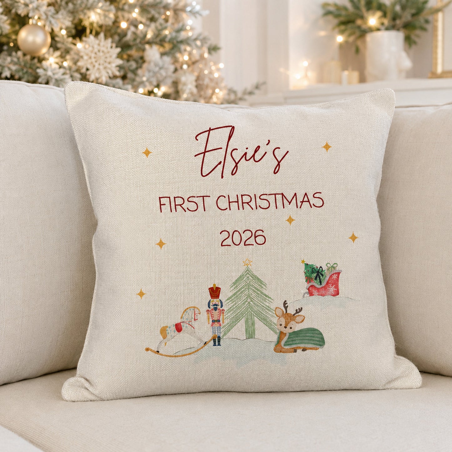 A personalized cream-colored throw pillow with embroidered text reading 'Elsie's FIRST CHRISTMAS 2026' and a festive Christmas illustration, resting on a light beige sofa with a blurred Christmas tree in the background.