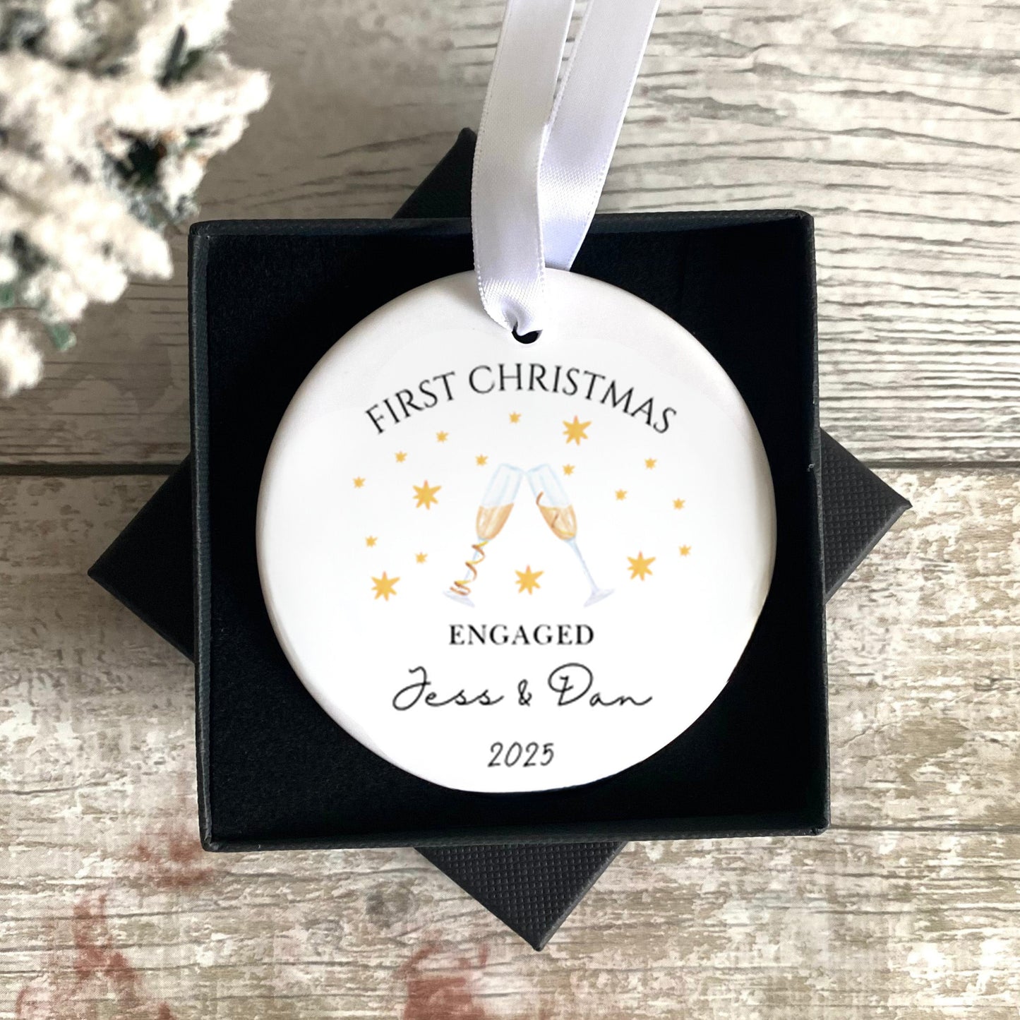 Personalised First Christmas Engaged ornament in a black box on a wooden surface with a snowy background.