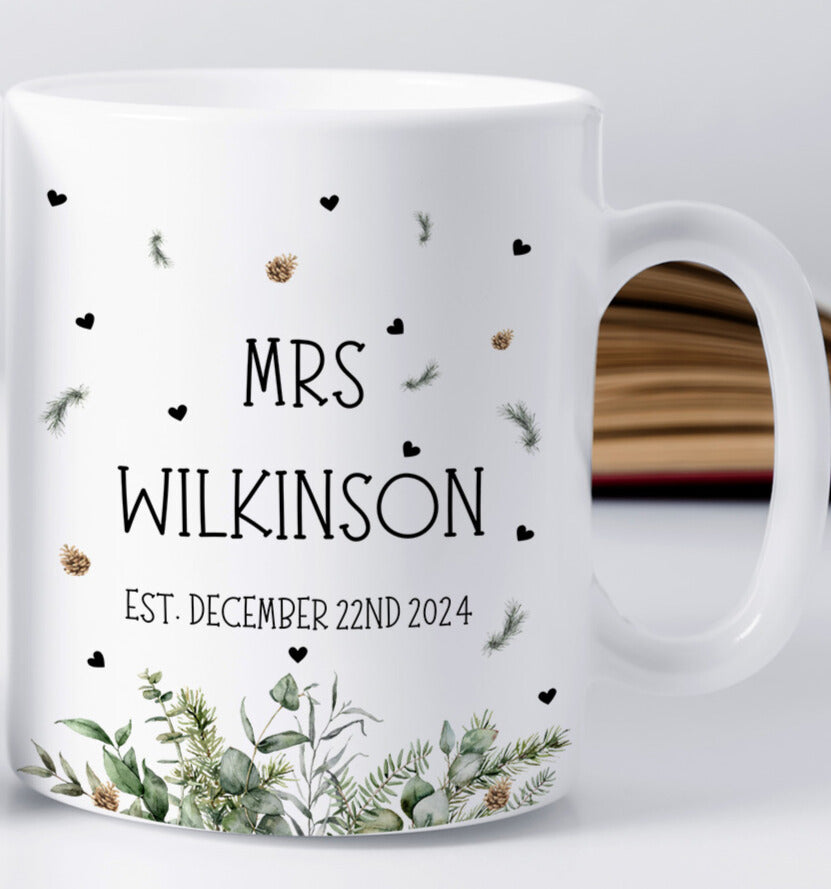 A white ceramic mug with the text "MRS WILKINSON" and "EST. DECEMBER 22ND 2024" printed in black capital letters. The mug is decorated with scattered black hearts, pine needles, and small gold pinecones. At the bottom, there is a detailed illustration of winter greenery with eucalyptus leaves and fir branches. A blurred open book is visible in the background.