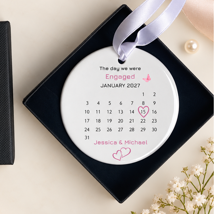 A personalized engagement ornament displayed next to its black presentation box on a styled flat-lay background with pink roses, baby's breath, pearls, and satin ribbons.