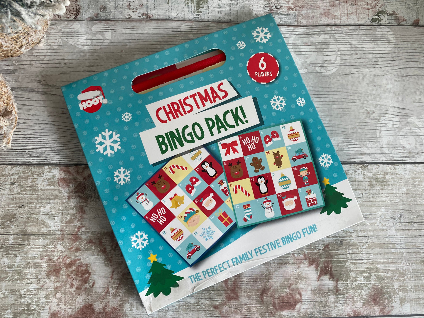 A festive Christmas Bingo Pack displayed on a rustic wooden background. The packaging is blue with white snowflakes and features the title “Christmas Bingo Pack!” in bold red, green, and white letters. Two colourful sample bingo cards with holiday-themed illustrations such as Santa, a reindeer, a snowman, and a Christmas tree are shown on the front. It is designed for 6 players. The bottom of the box reads, “The perfect family festive bingo fun!” making it a cheerful activity for holi