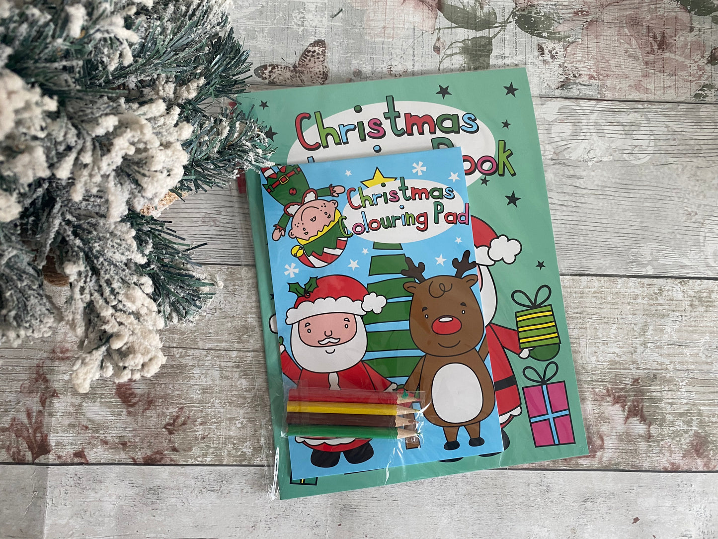 Children’s Christmas colouring book and pad set with festive Santa and reindeer illustrations, includes colouring pencils, displayed on a rustic wooden background with snowy tree decor.