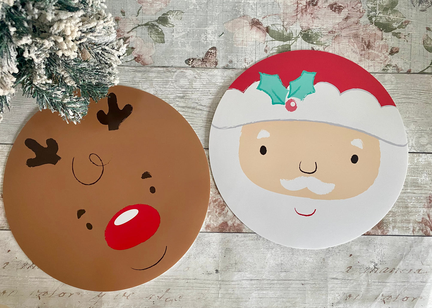 Two round, festive placemats featuring adorable character designs. One mat shows Rudolph the Red-Nosed Reindeer with a red nose, curved smile, and antlers on a brown background. The second mat displays Santa Claus with rosy cheeks, a white beard, and a red hat with holly detail, set against a white background. Perfect for Christmas table settings.