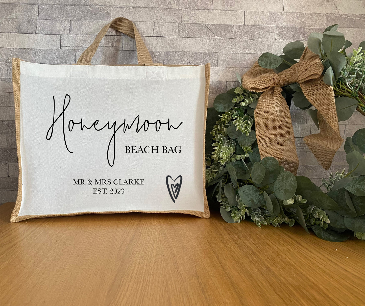 A personalized honeymoon beach bag made of canvas and jute, with the words "Honeymoon Beach Bag" in cursive and "MR & MRS CLARKE EST. 2023" in smaller, serif font. The bag is adorned with a heart symbol and placed on a wooden surface beside a green plant with a decorative burlap bow.