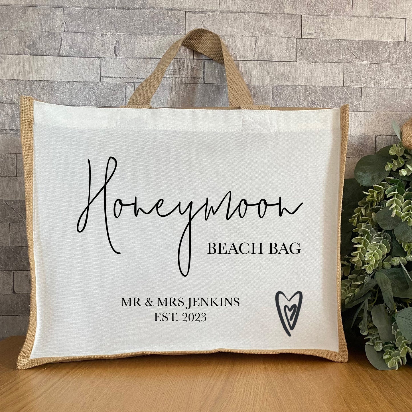 A jute and canvas beach bag with the words “Honeymoon Beach Bag” printed in elegant cursive, and “MR & MRS JENKINS EST. 2023” in a smaller, serif font. The bag has a heart design and is positioned on a wooden surface beside a bunch of green leaves.