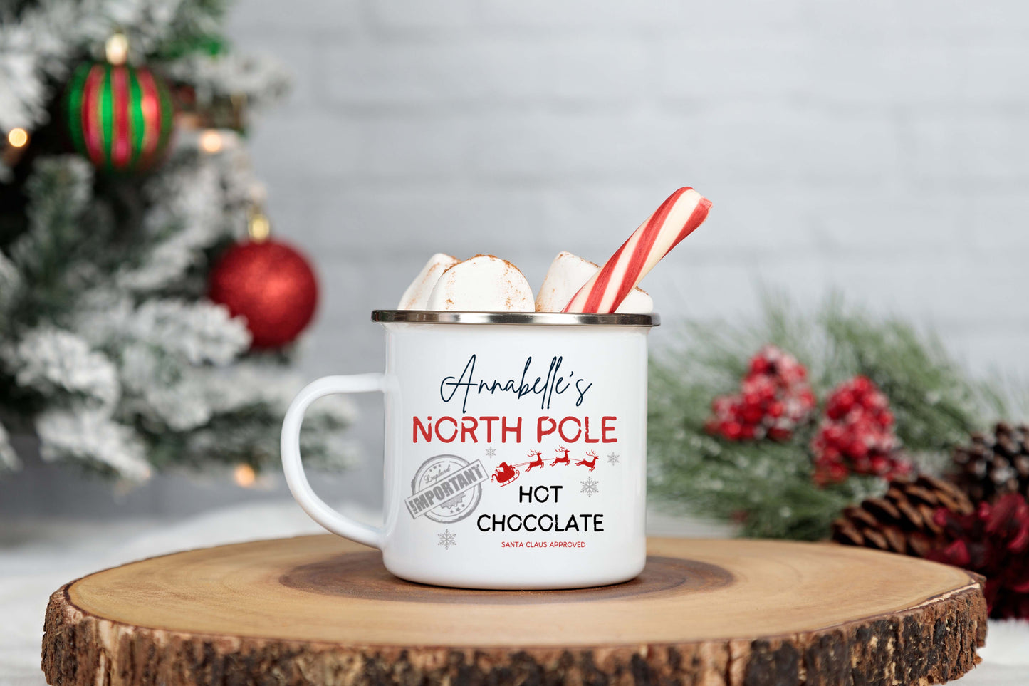 White enamel mug with 'North Pole Hot Chocolate' text, surrounded by Christmas decorations on a wooden surface.