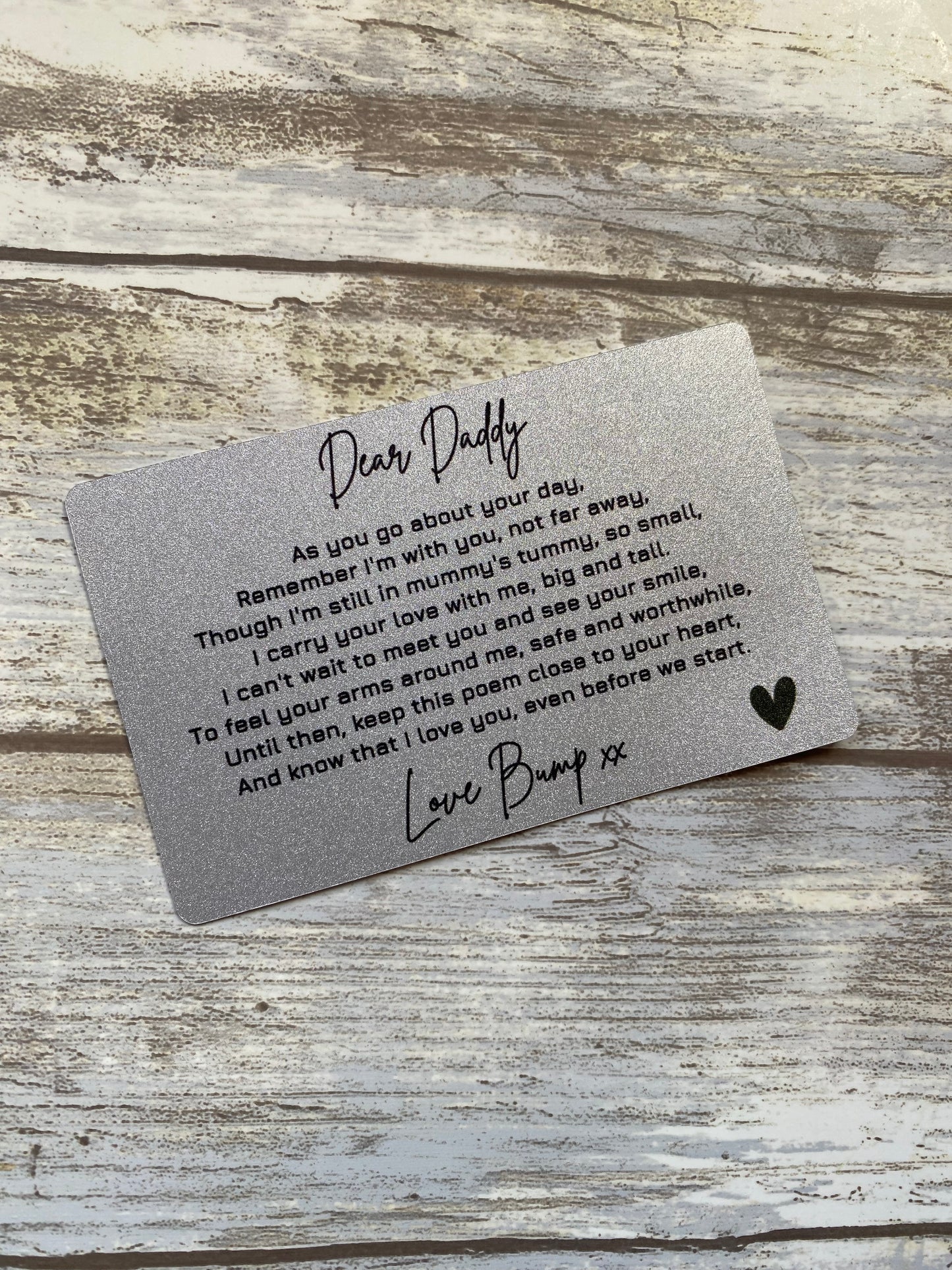 A silver wallet-sized card placed on a rustic wooden surface. The card contains a heartfelt message to a father from an unborn child, expressing love and anticipation for their first meeting. The text is printed in black script and regular font, ending with "Love Bump xx" and a heart symbol.