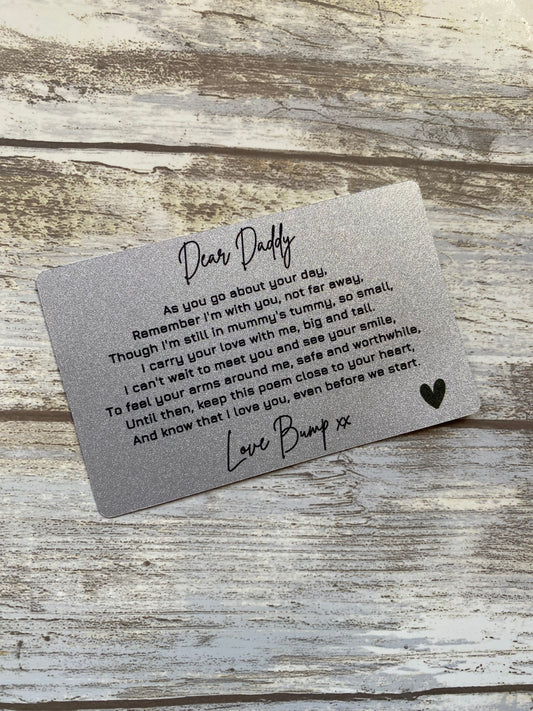 A silver wallet-sized card placed on a rustic wooden surface. The card contains a heartfelt message to a father from an unborn child, expressing love and anticipation for their first meeting. The text is printed in black script and regular font, ending with "Love Bump xx" and a heart symbol.