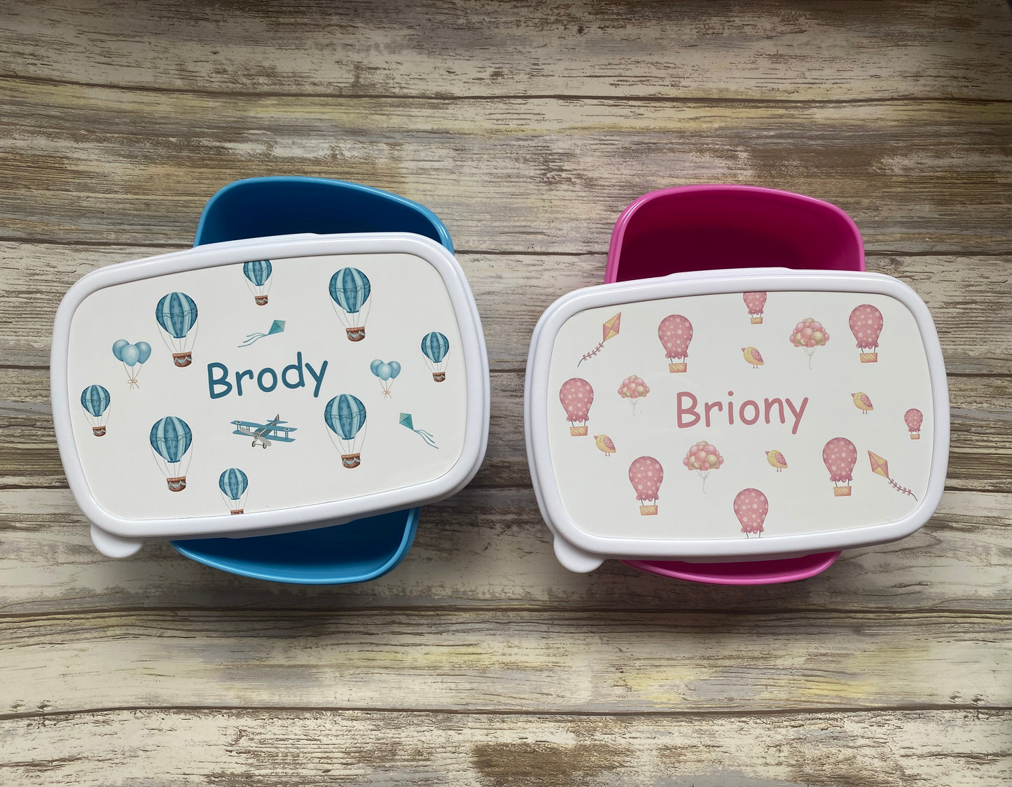 Set of unique personalised lunch boxes with colourful designs. Blue lunch box featuring hot air balloons and an airplane for ‘Brody’, and pink lunch box with hot air balloons and birds for ‘Briony’. Perfect gift for kids’ school or outings.
