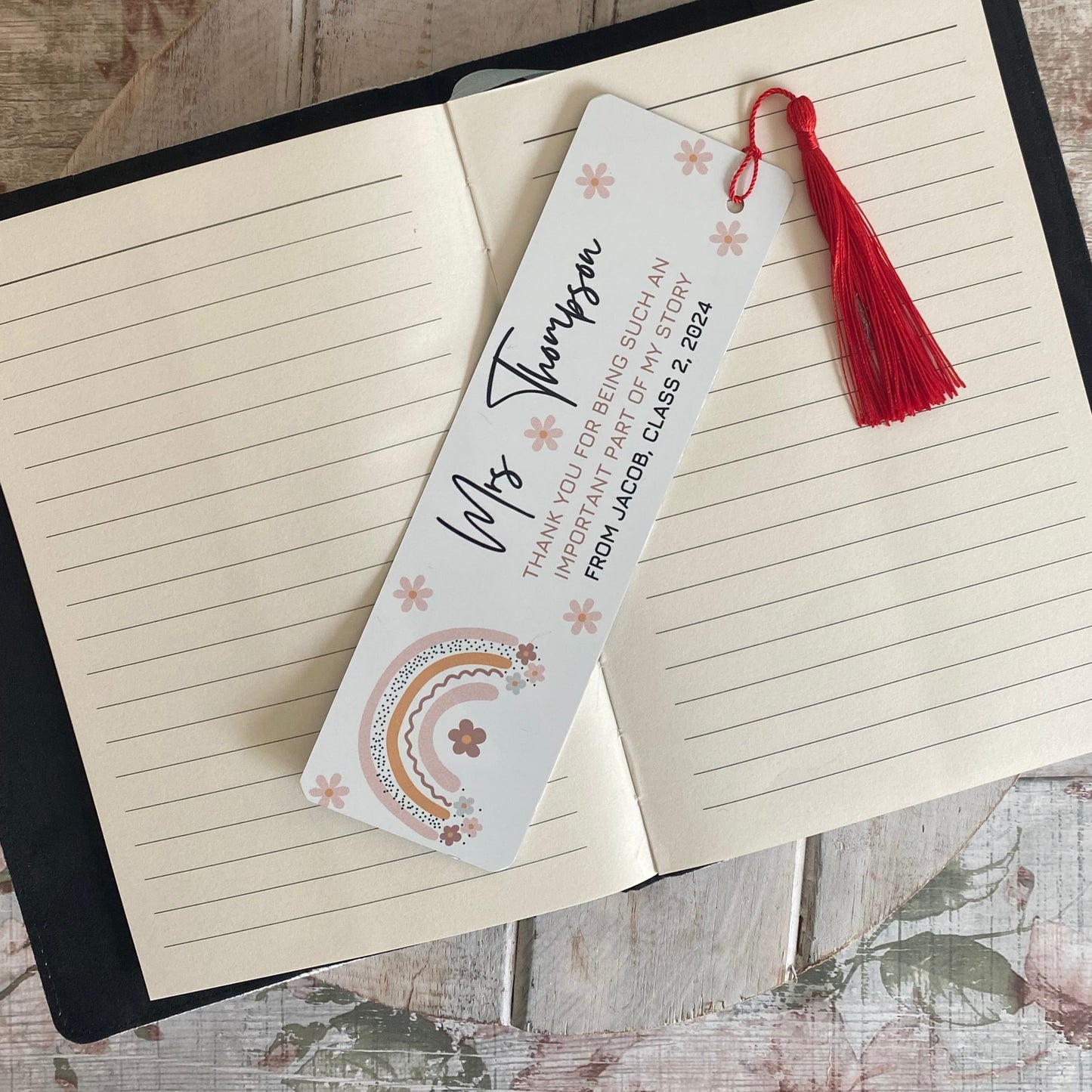 Personalised teacher bookmark with red tassel, featuring rainbow and floral design – thank you gift for Miss Thompson from a student, displayed in a lined notebook.