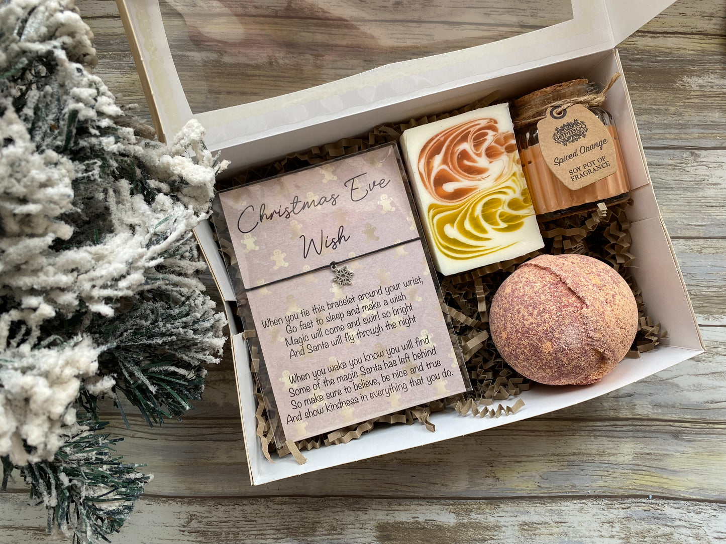 Gift box with Christmas Eve Wish card, soap, and bath bomb on a wooden surface with a decorative tree.