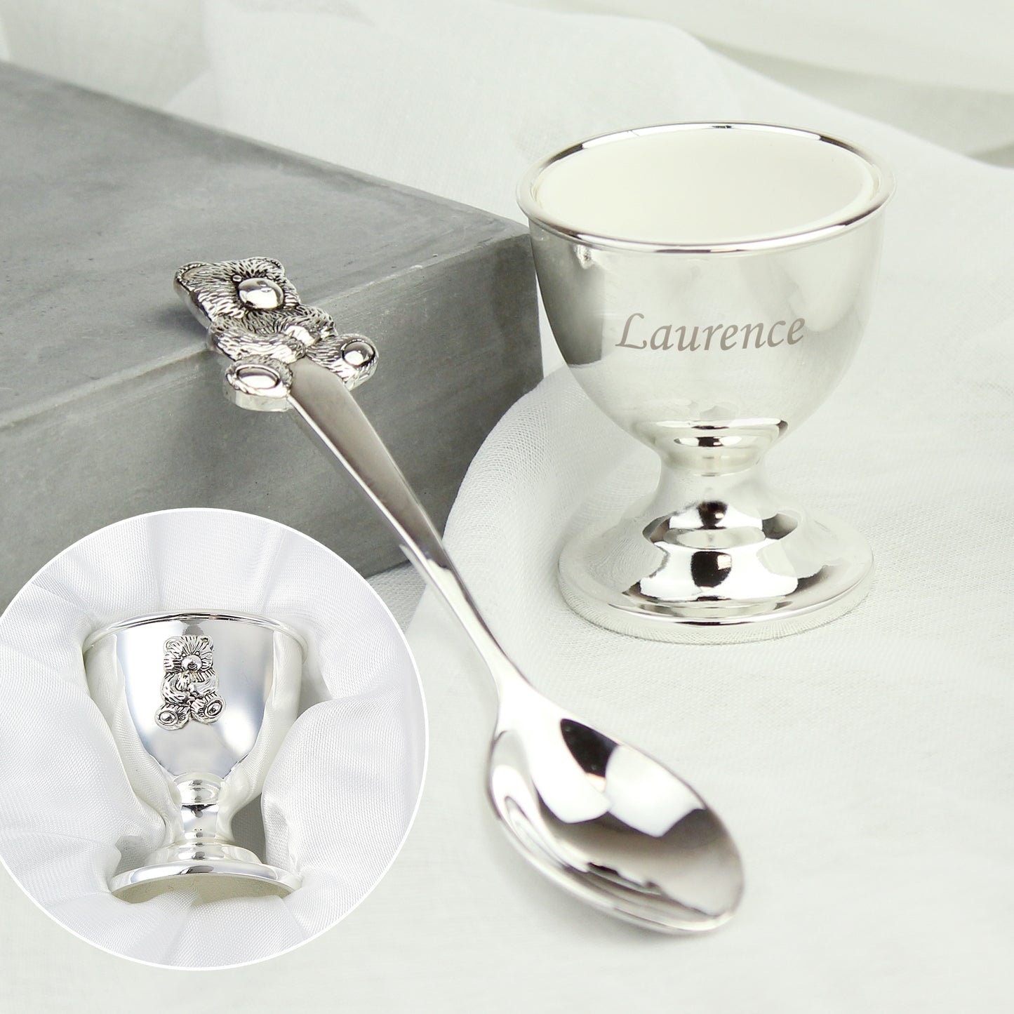 Silver spoon and egg cup set with 'Laurence' on a white surface.
