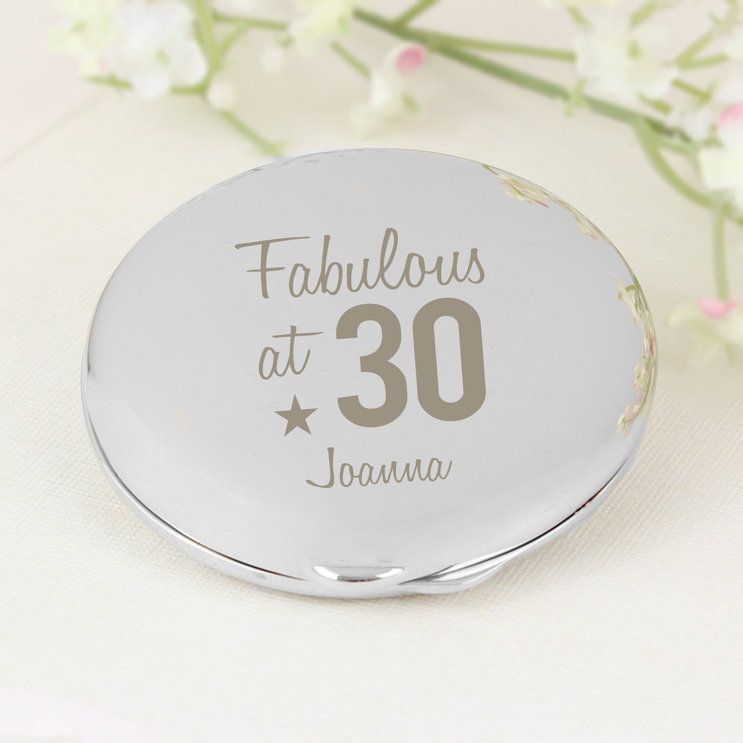 Round silver compact mirror with 'Fabulous at 30 Joanna' text on a light background with flowers.