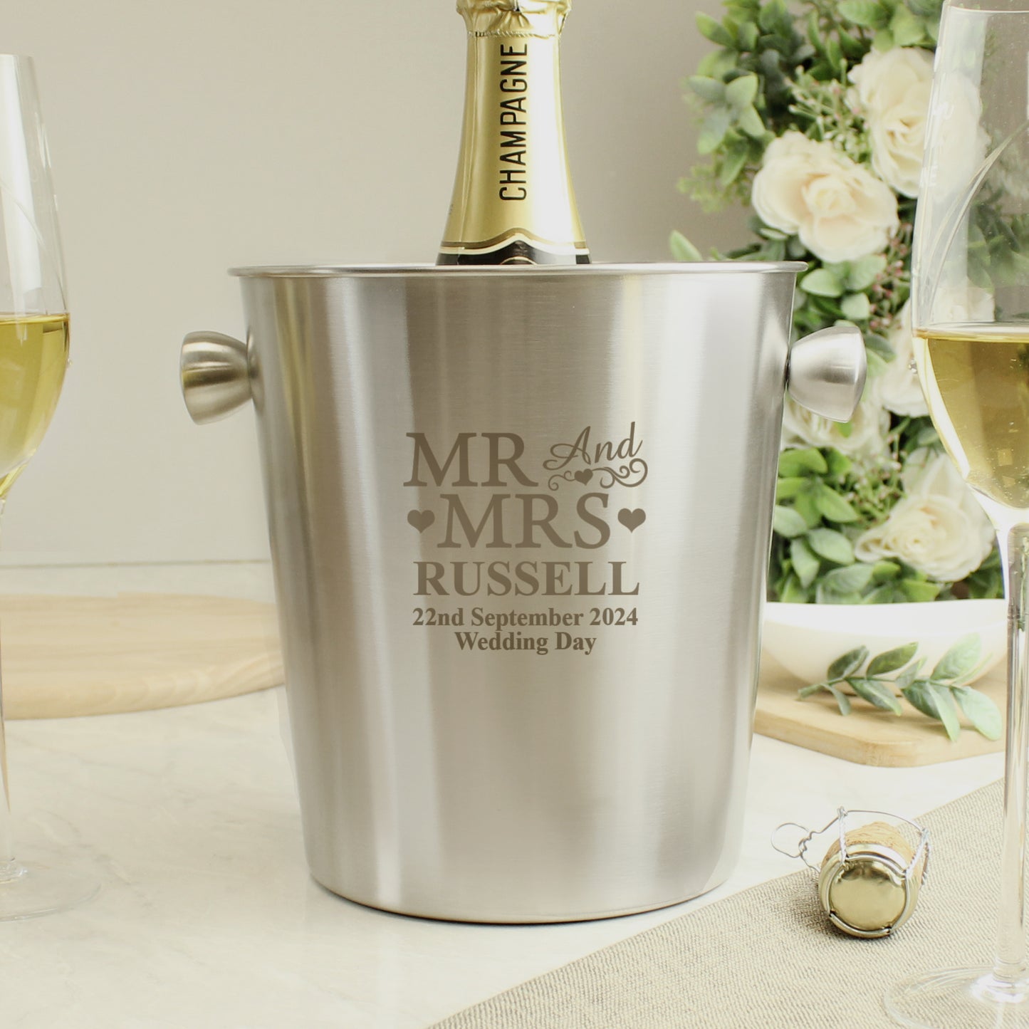 Stainless steel ice bucket engraved with 'MR & MRS RUSSELL' and wedding details, surrounded by champagne and glasses.