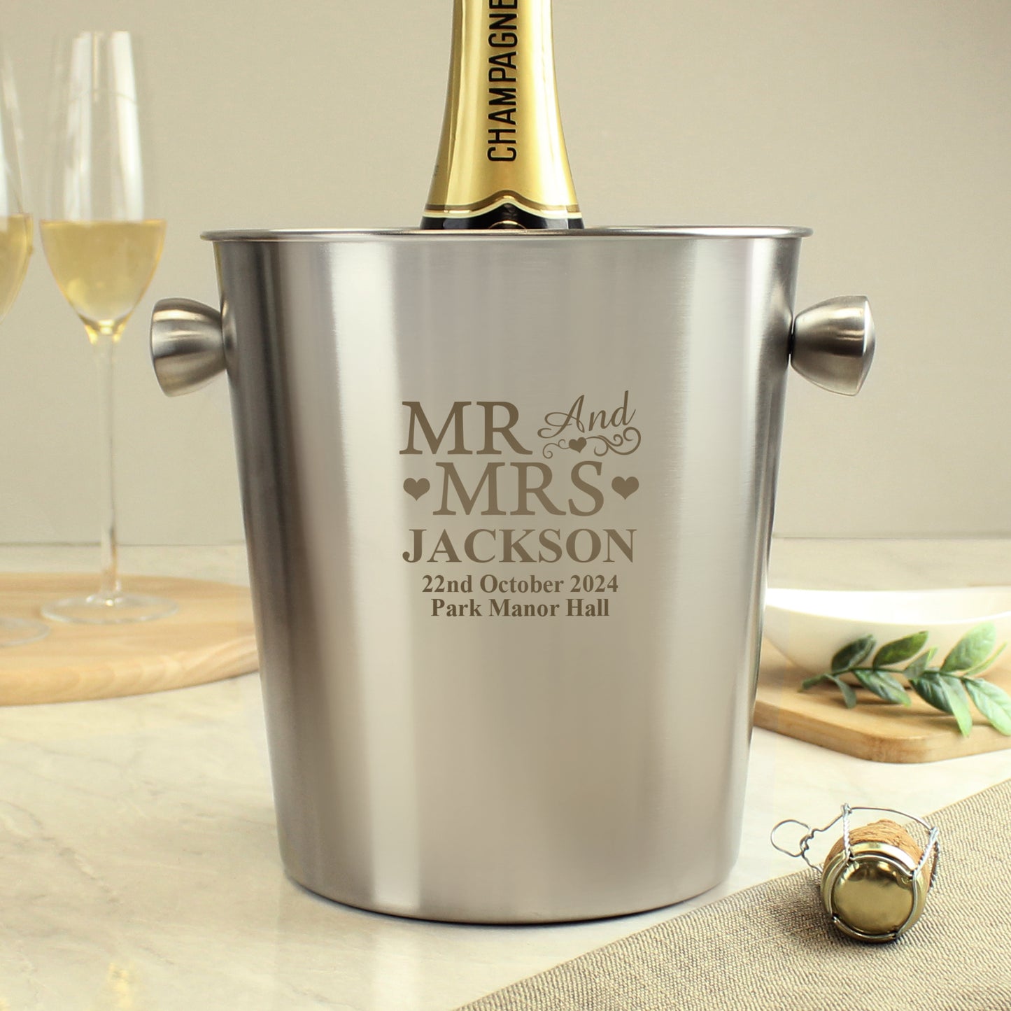 Personalised champagne bucket with engraved text on a table with glasses and a bottle cap.