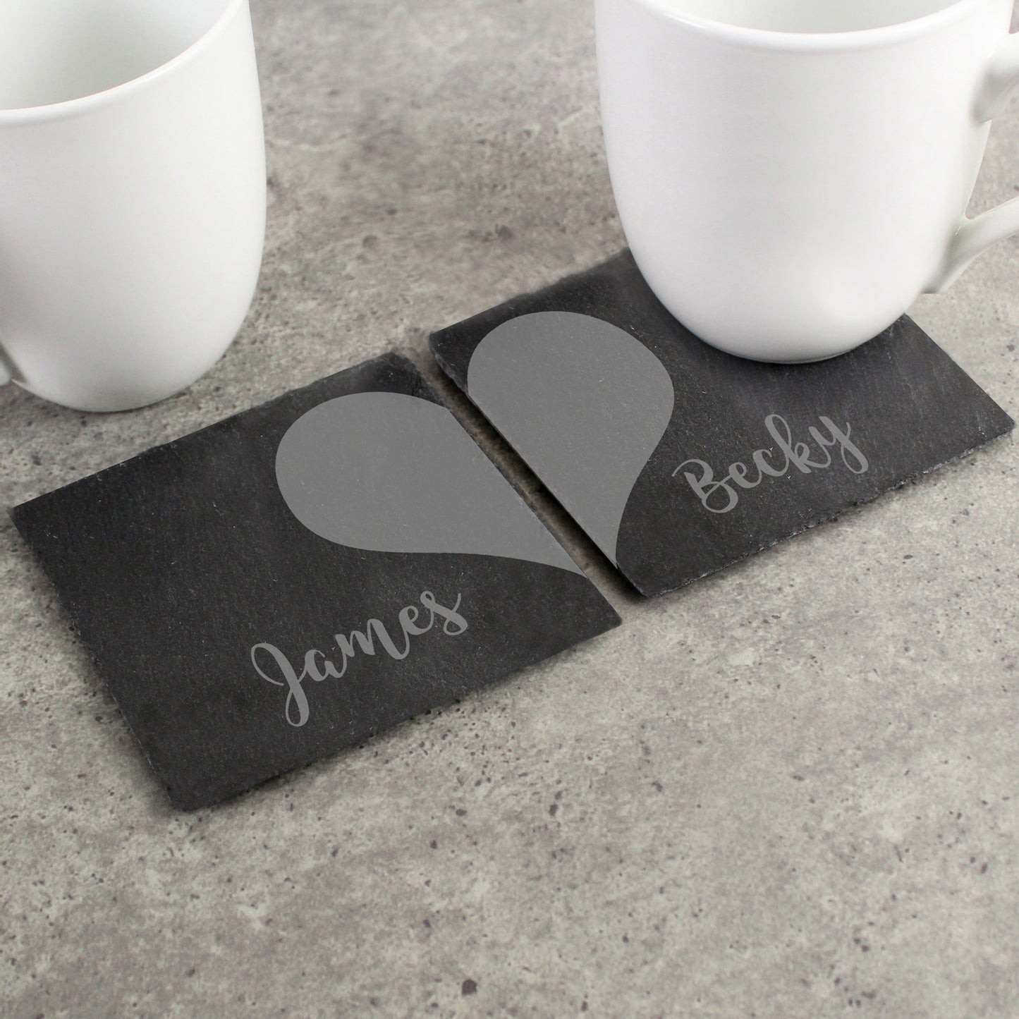 Two slate coasters with grey heart designs and names 'James' and 'Becky' on a concrete surface.