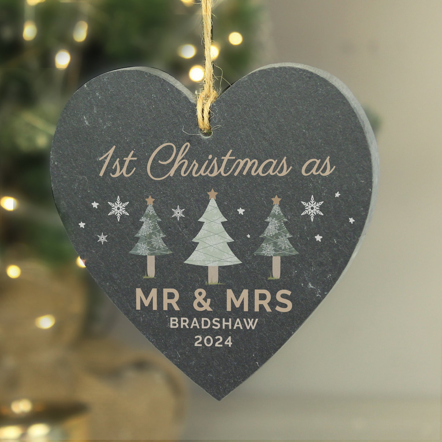 Heart-shaped Christmas ornament with '1st Christmas as Mr & Mrs Bradshaw 2024' text, trees, and snowflakes against a blurred Christmas tree background.