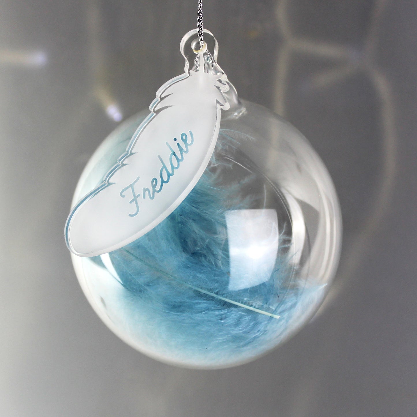 Decorative ornament with blue feathers inside a clear sphere and a name tag on a dark background