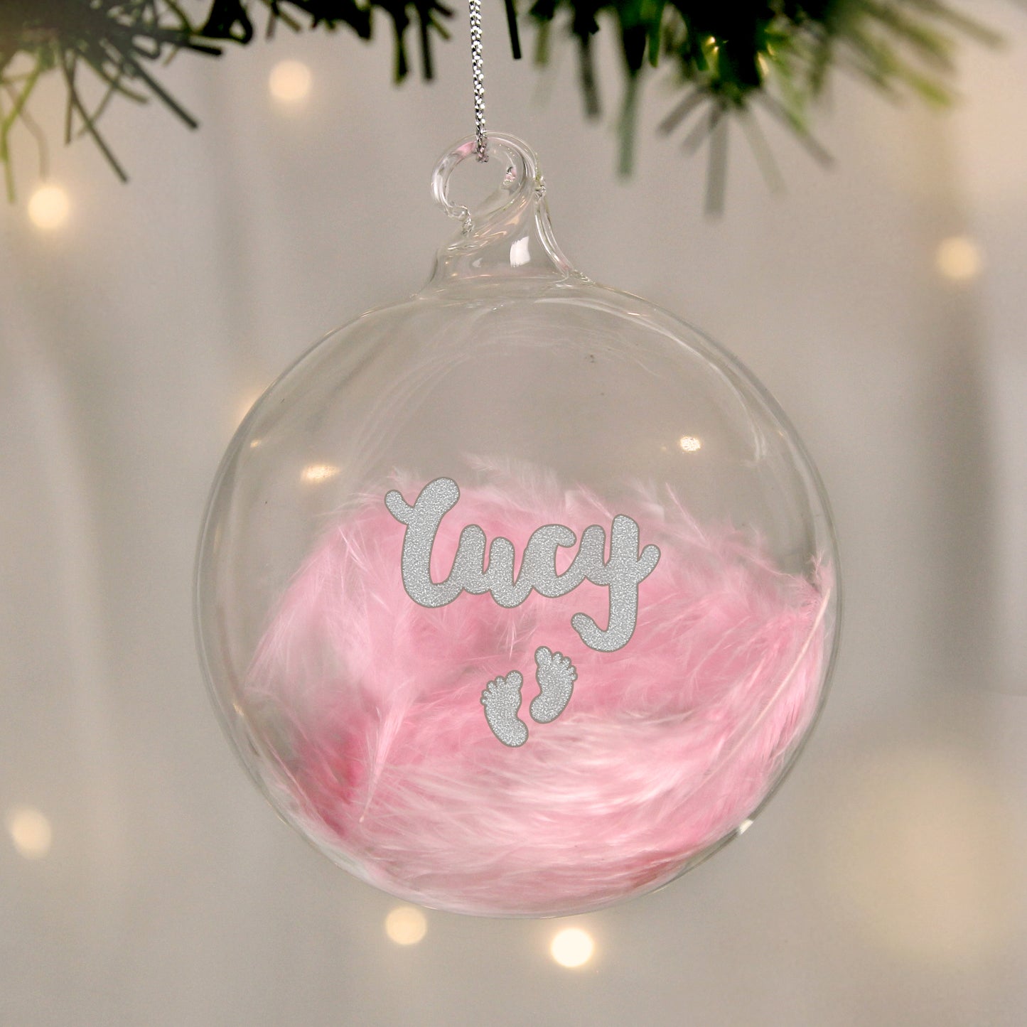 A clear glass bauble filled with soft pink feathers, personalized with the name “Lucy” in silver glittery text. Below the name are two small silver glitter baby footprints. The bauble is hanging from a silver string on a softly lit Christmas tree, creating a warm and festive atmosphere perfect for a baby girl's first holiday.