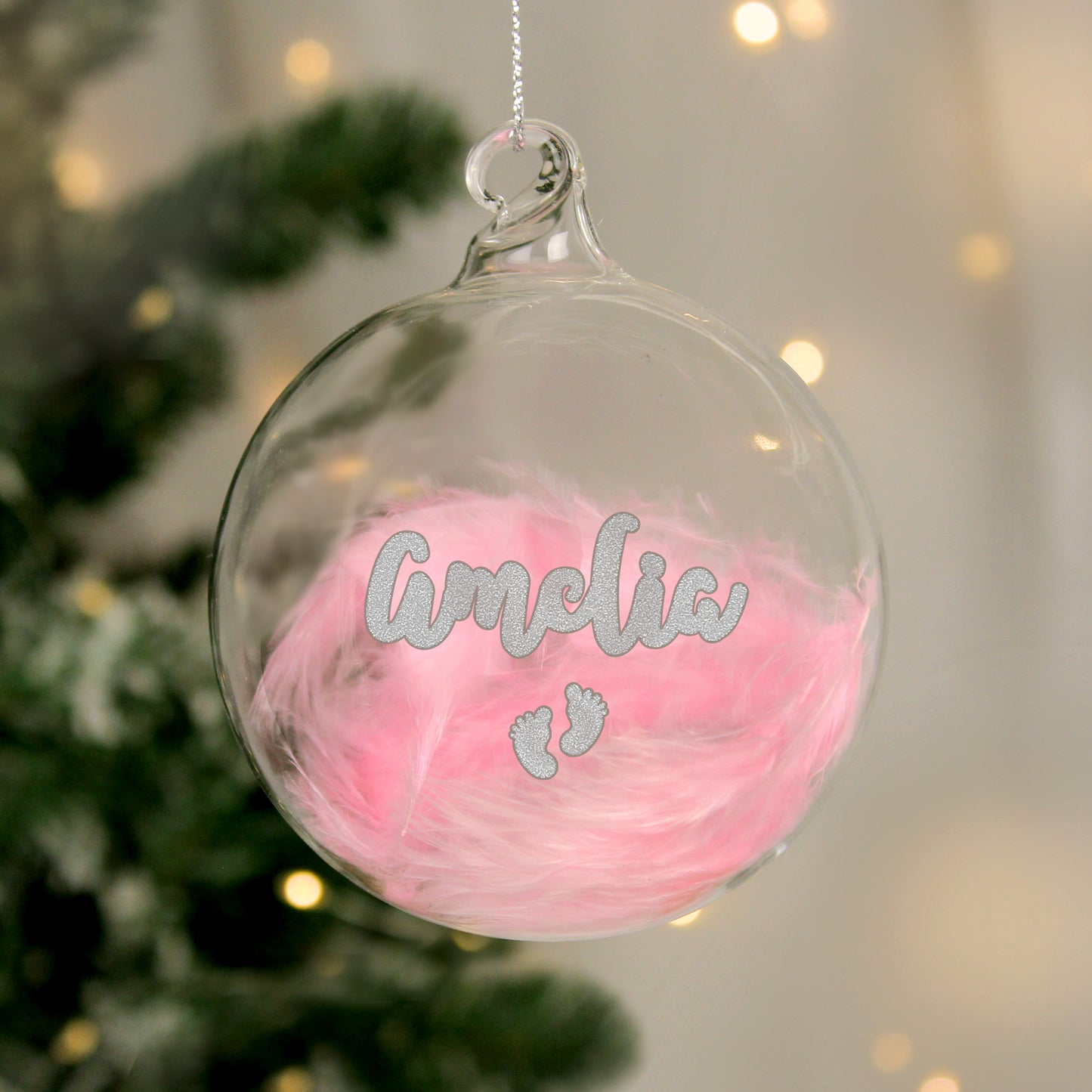 Personalised clear glass bauble filled with soft pink feathers, featuring the name “Amelia” in silver glitter script above a pair of tiny glitter baby footprints. A sweet and sentimental decoration for a baby girl's first Christmas.