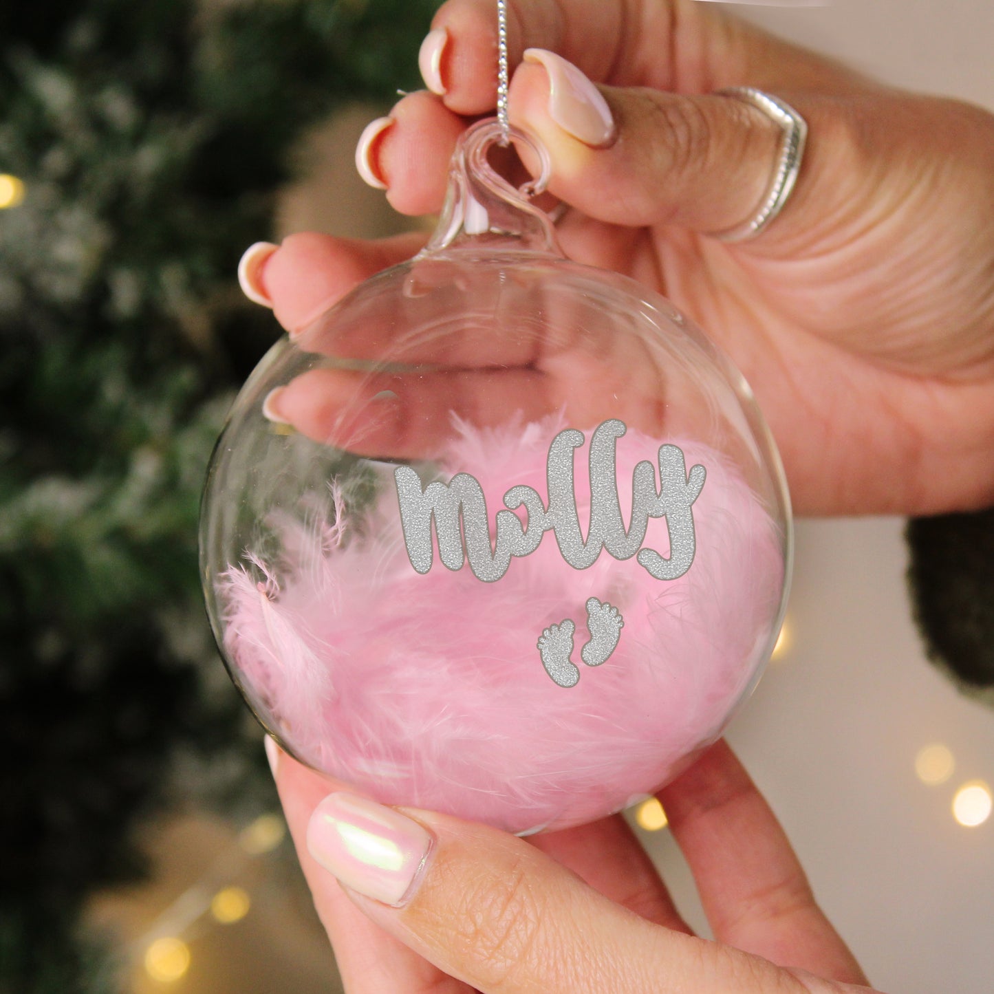 Clear glass bauble filled with delicate pink feathers, featuring the name “Molly” in sparkling silver glitter text. Below the name are two small silver glitter baby footprints. The bauble is being held up by a hand, showcasing a soft and sentimental design ideal for celebrating a baby girl's first Christmas.