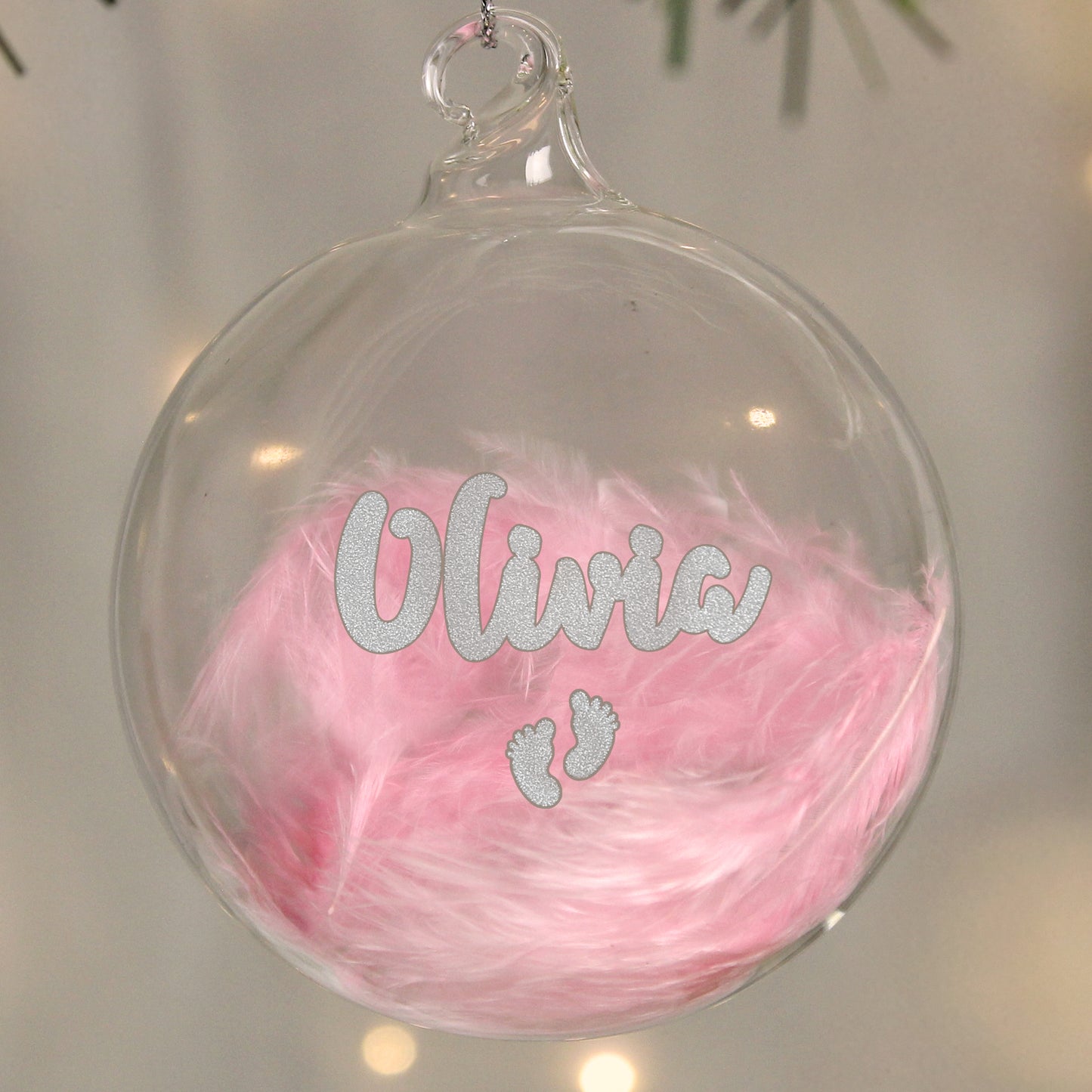 Personalised glass plastic bauble containing soft pink feathers, decorated with the name “Olivia” in sparkling silver glitter script. Below the name are two tiny glitter baby footprints. A sweet and sentimental ornament, perfect for a baby girl's first Christmas.