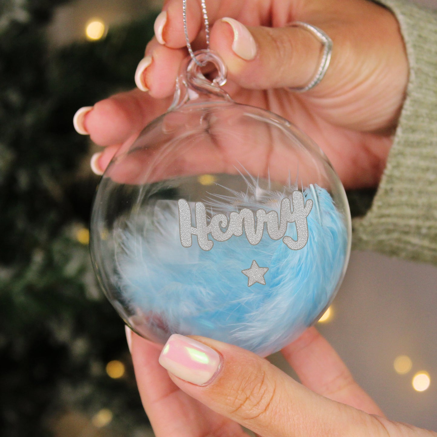 Personalised glass bauble filled with soft blue feathers, featuring the name “Henry” in silver glitter text above a glitter star. Held gently in hand, this custom baby boy ornament makes a sweet festive keepsake.