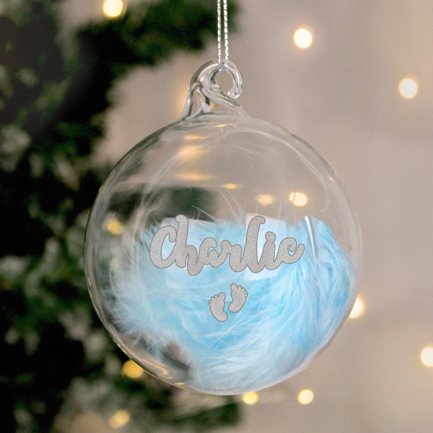 Clear plastic Christmas bauble filled with soft blue feathers, personalised with the name “Charlie” and silver glitter baby footprints. A charming, custom decoration perfect for celebrating a baby boy’s first Christmas or as a keepsake gift.