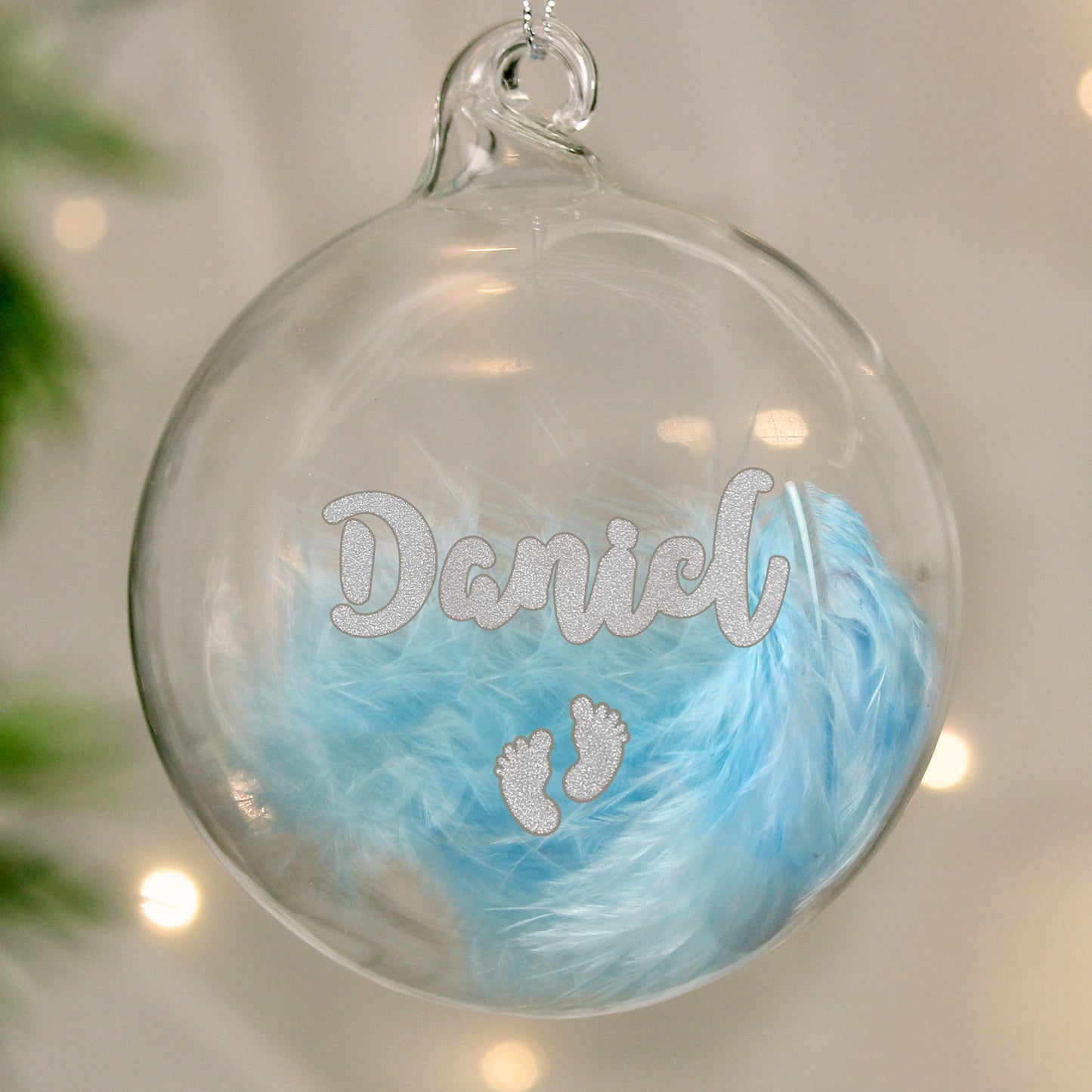 Customisable clear plastic Christmas bauble filled with soft blue feathers, featuring glittery silver text reading "Daniel" and a pair of baby footprints. A beautiful personalised decoration perfect for celebrating a baby's first Christmas or as a cherished keepsake.