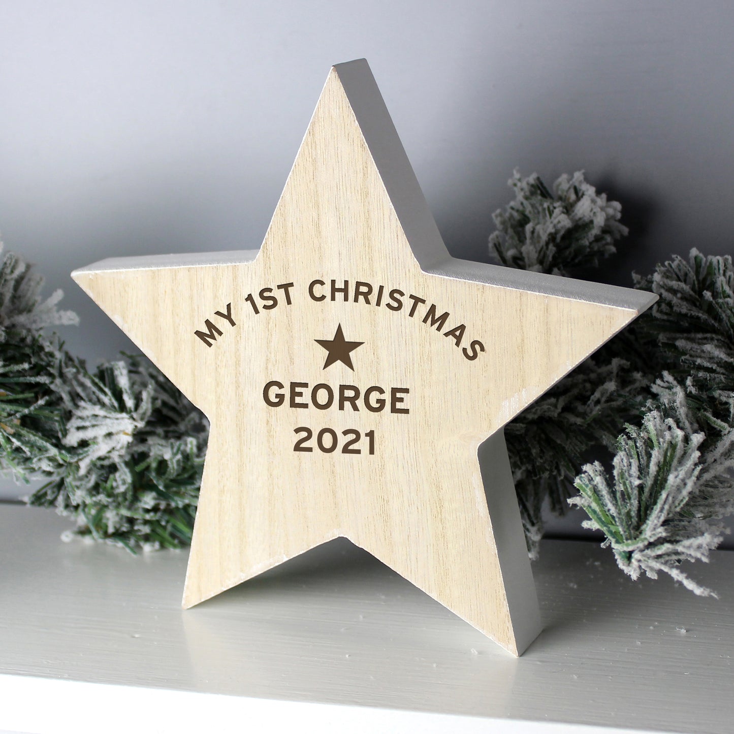 Personalised Wooden star ornament with 'My 1st Christmas George 2021' engraving, surrounded by festive greenery.