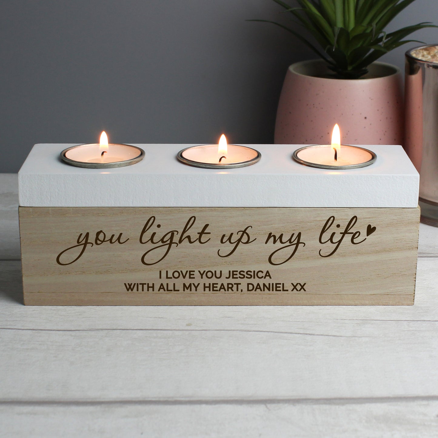 Three tealight holder with engraved message on a wooden base, placed on a surface with a plant in the background.
