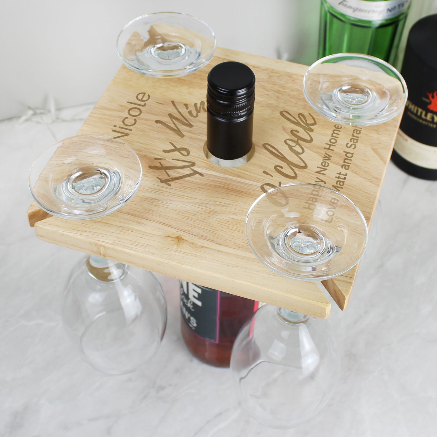 Wooden tray with engraved text holding four wine glasses and a bottle on a marble surface.