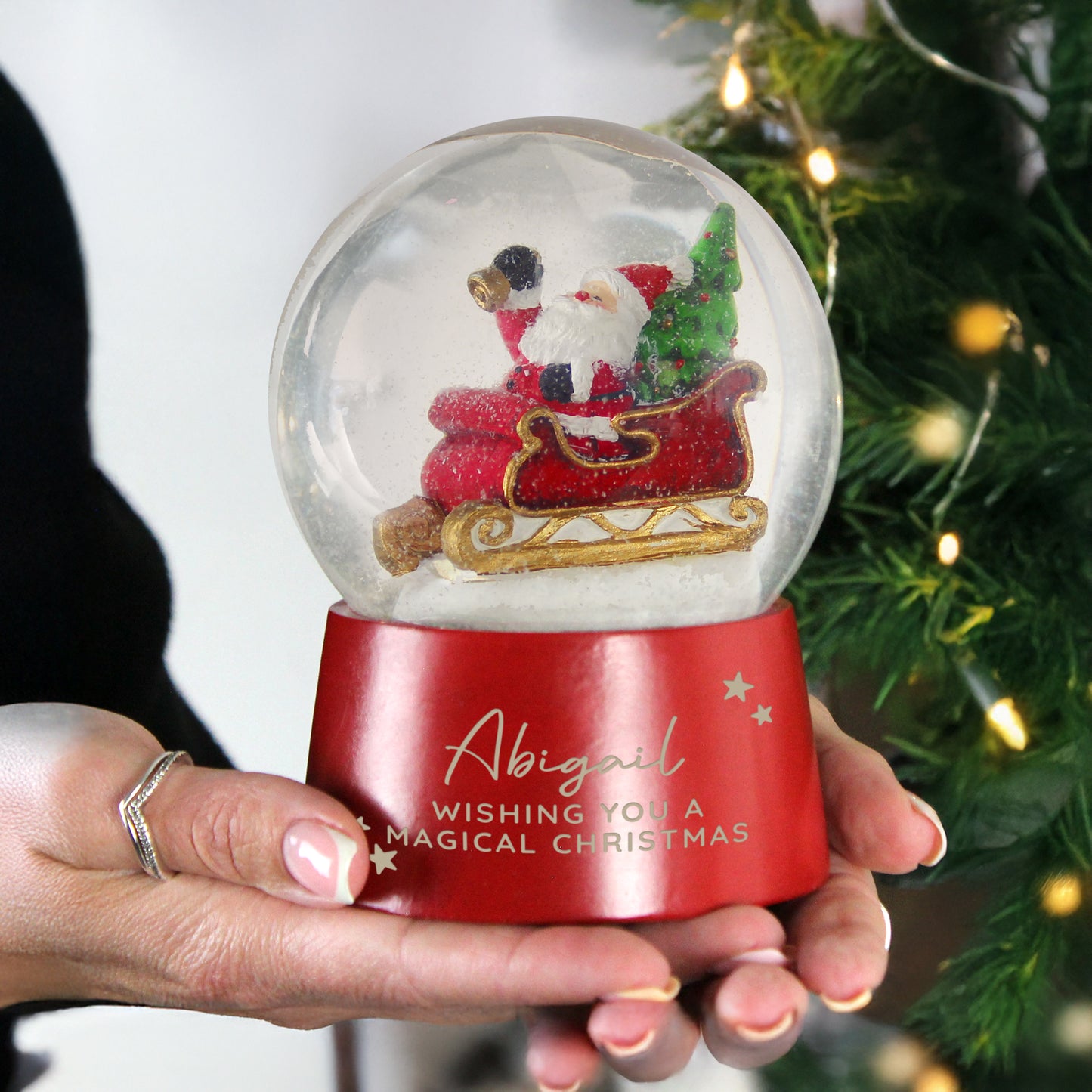 Hand holding a Christmas snow globe with Santa Claus and a tree, against a blurred Christmas tree background.
