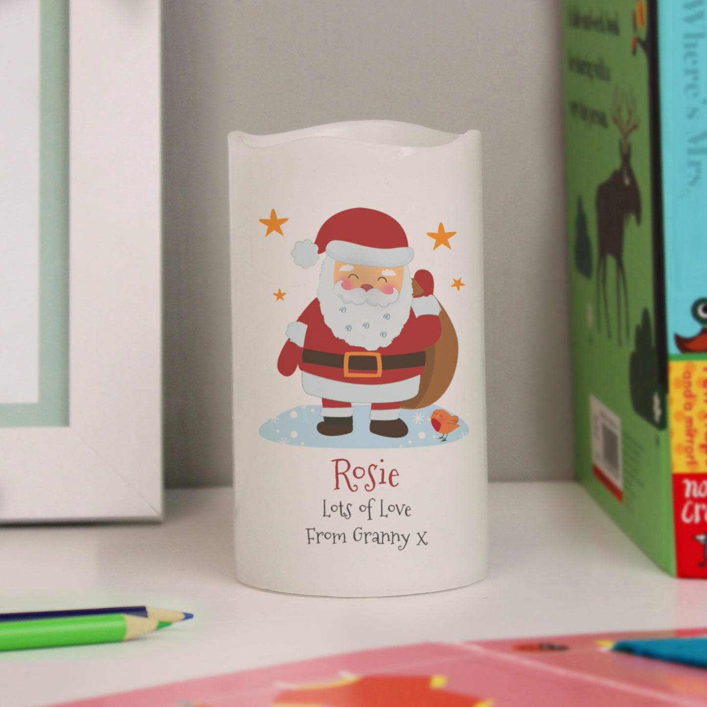 Festive LED candle with Santa Claus design on a white surface with Christmas-themed books in the background.