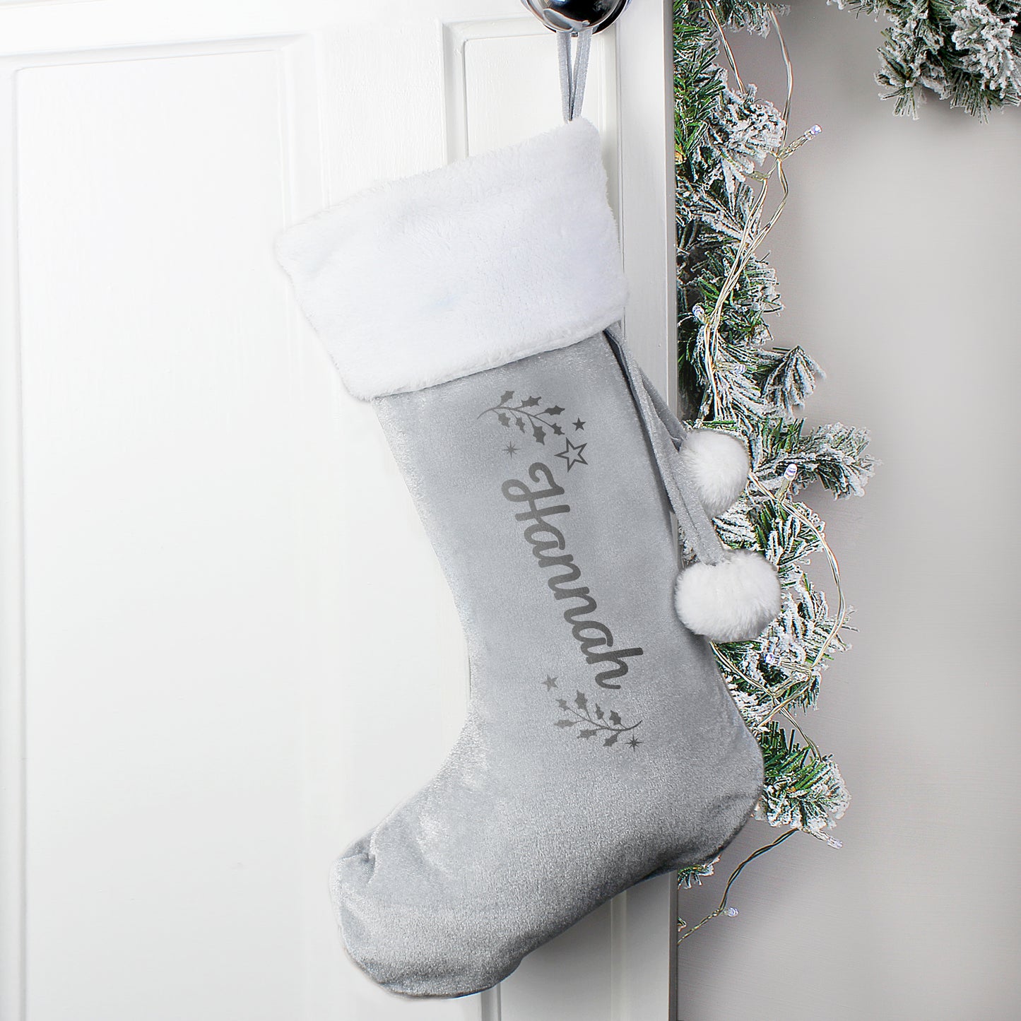 Gray Christmas stocking with white pom-poms hanging on a white door with decorative greenery.