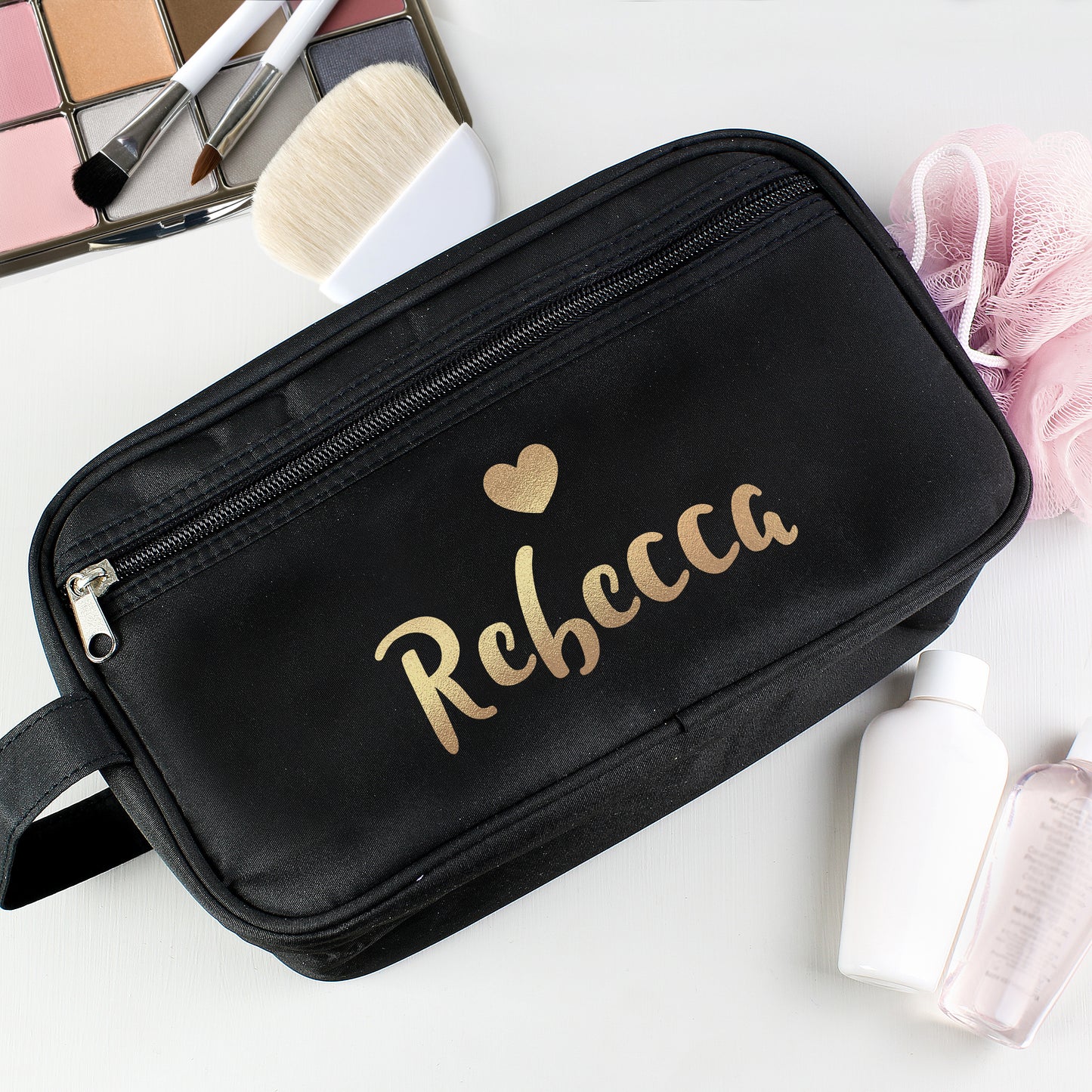 Black makeup bag with 'Rebecca' and a heart design on a white surface with makeup items.