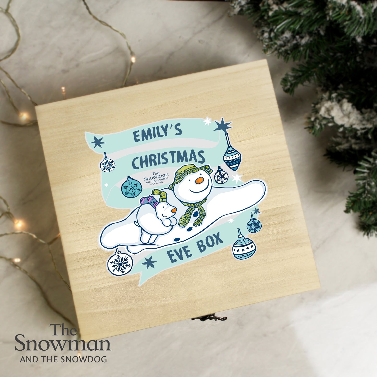 Wooden box with 'Emily's Christmas Eve Box' design featuring snowmen and festive elements, surrounded by Christmas decorations.