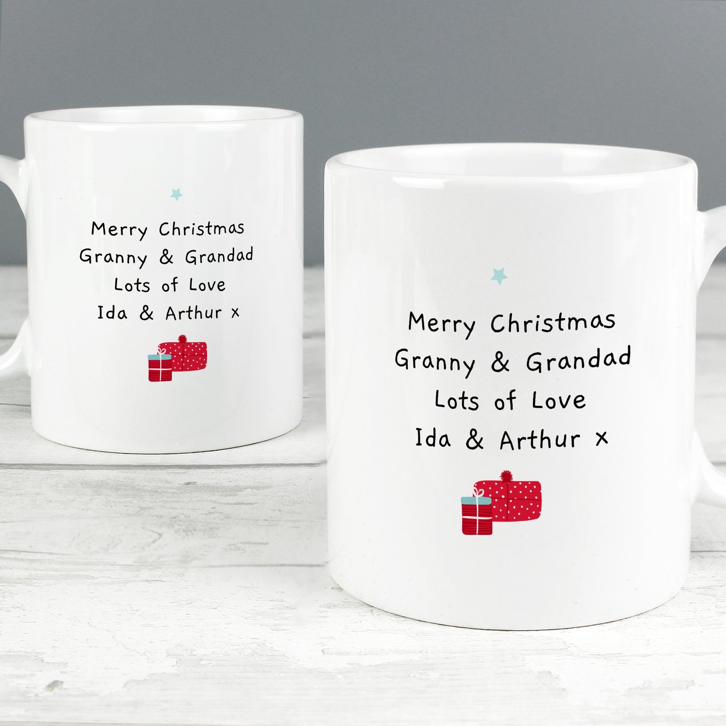 Two white mugs with Christmas-themed text and gift icons on a wooden surface.