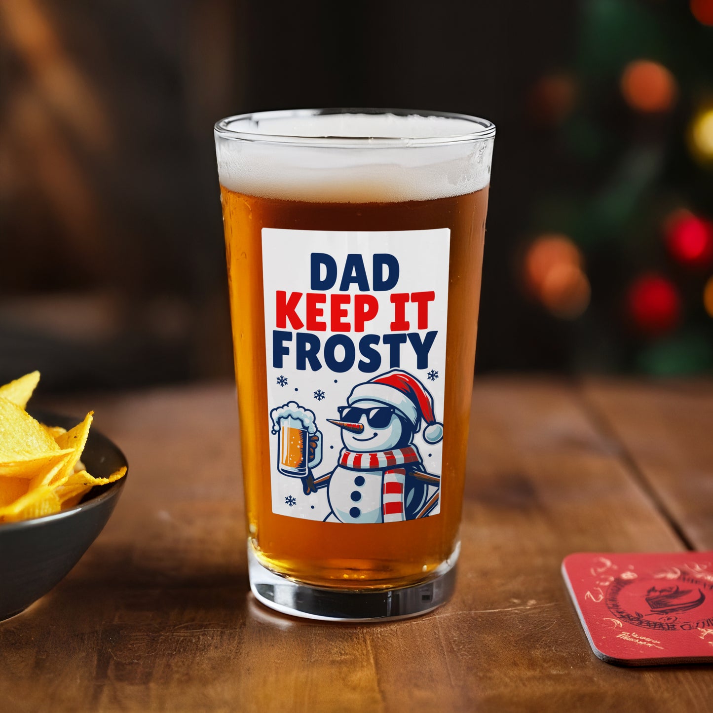 Personalised Beer glass with 'Dad Keep It Frosty' label on a wooden table with chips and a coaster.