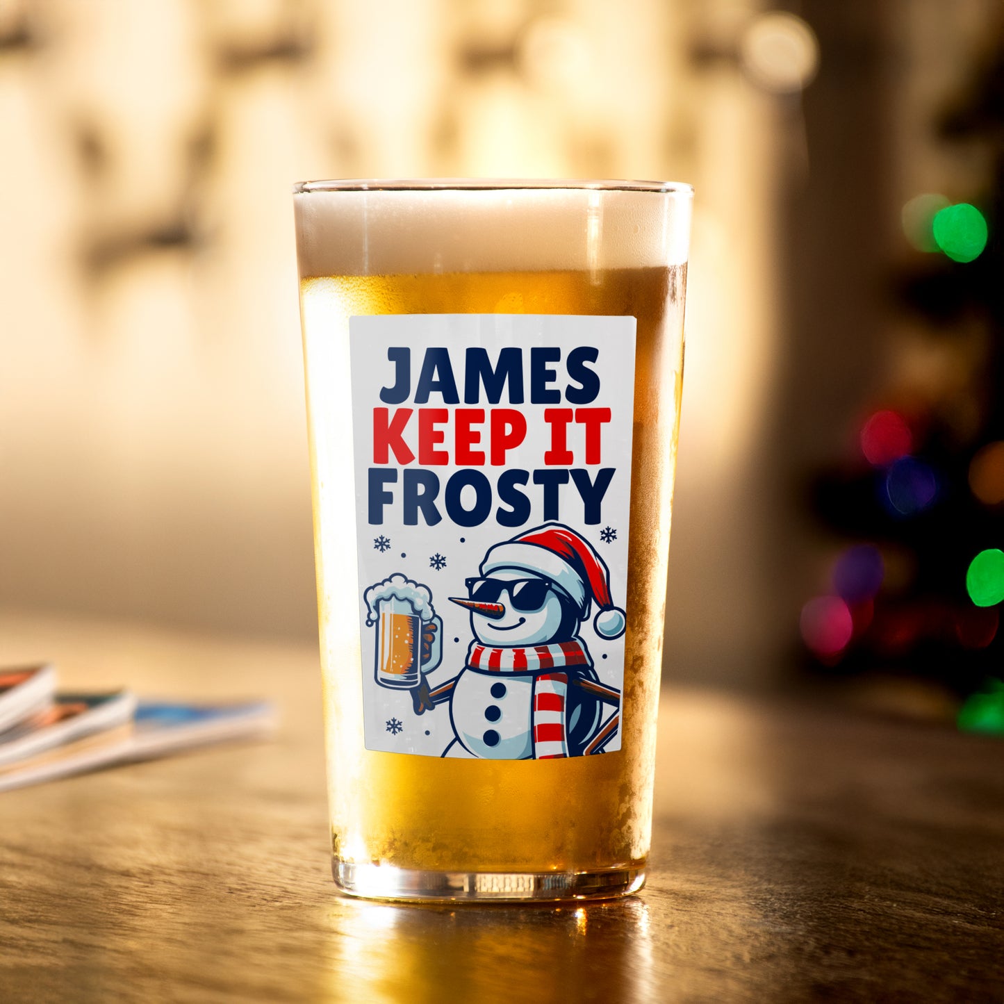 Pint glass with a snowman design and text on a wooden surface with blurred lights in the background