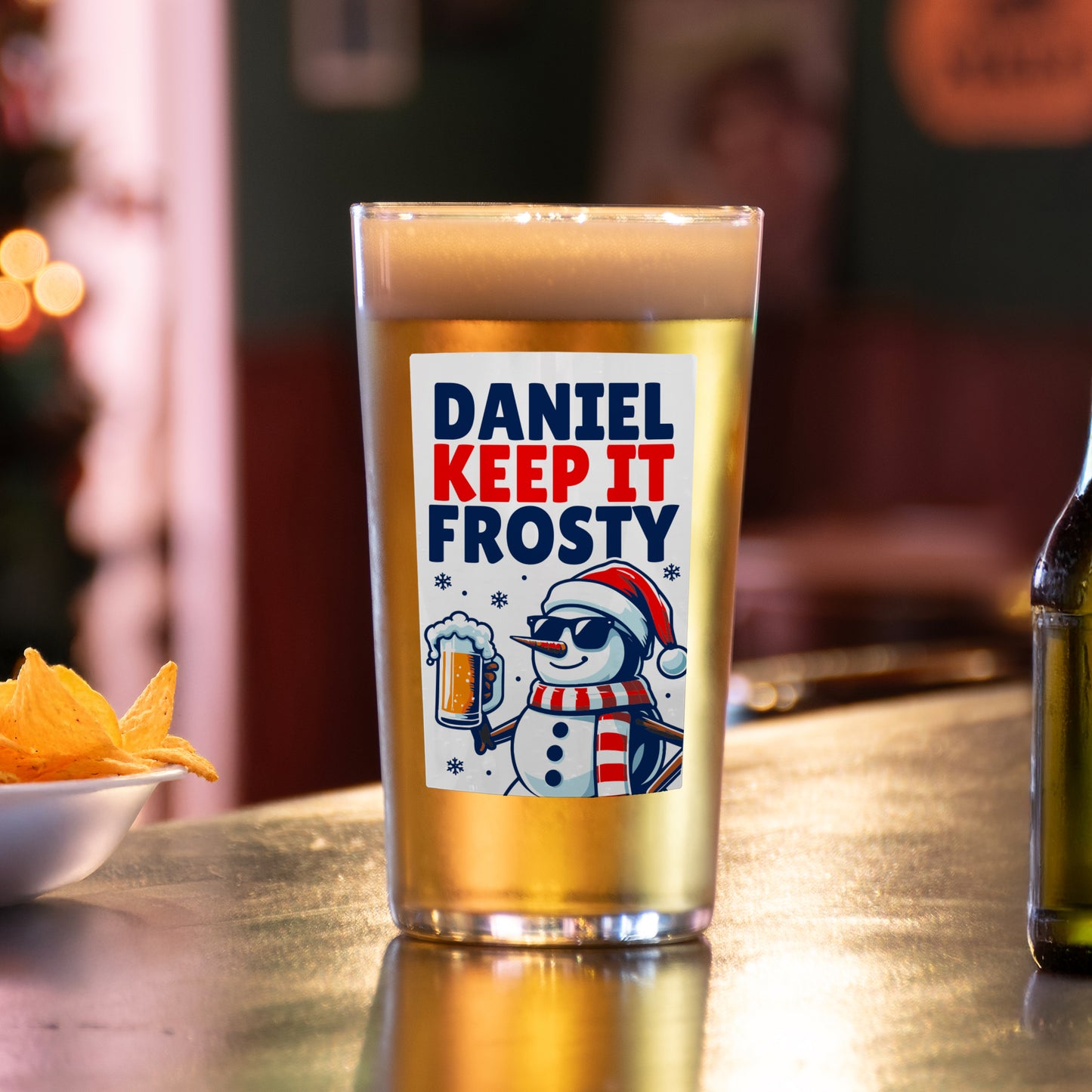 Beer glass with a 'Daniel Keep It Frosty' label on a bar counter with snacks and a bottle.