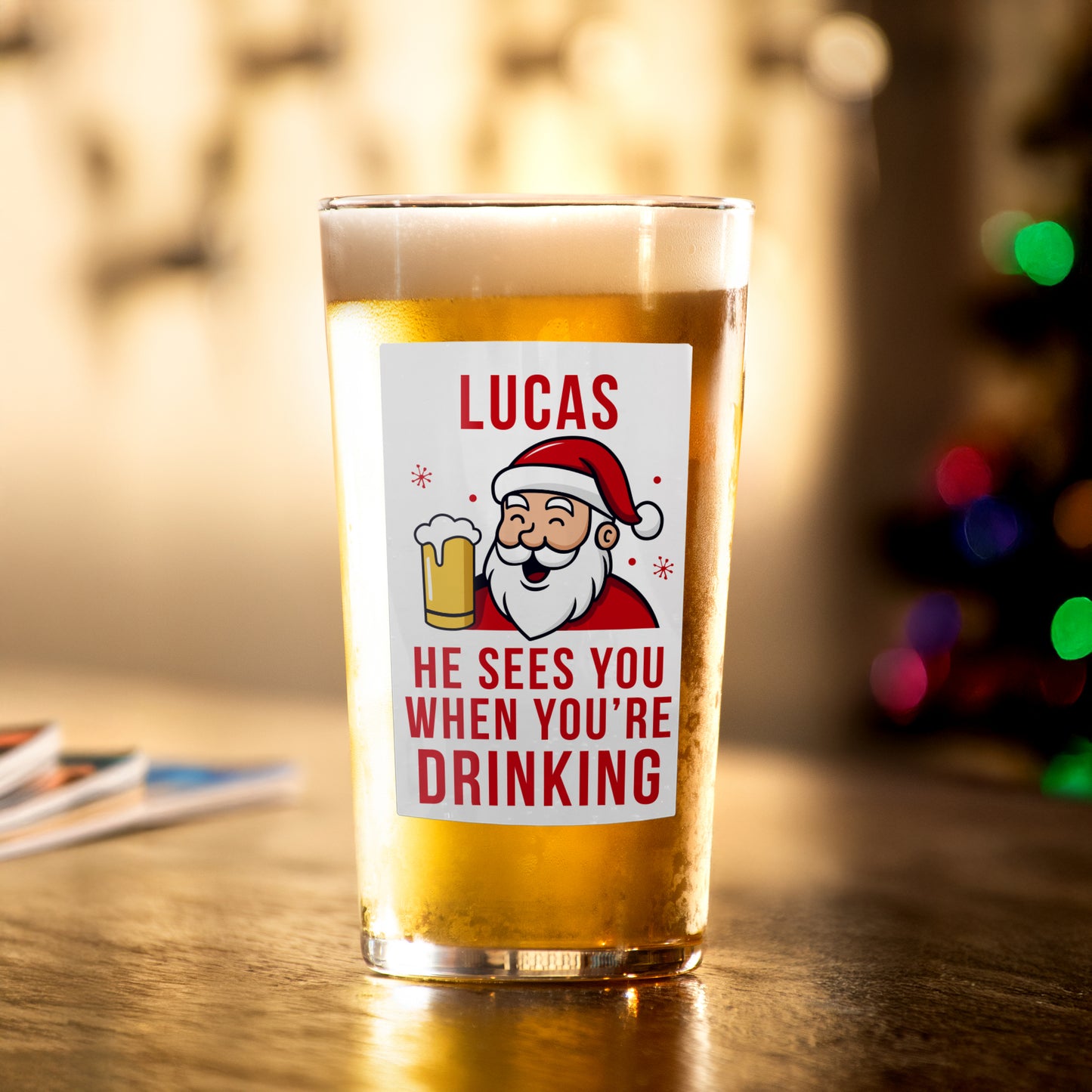 Personalised Beer glass with Santa Claus label and text 'Lucas sees you when you're drinking' on a wooden table.