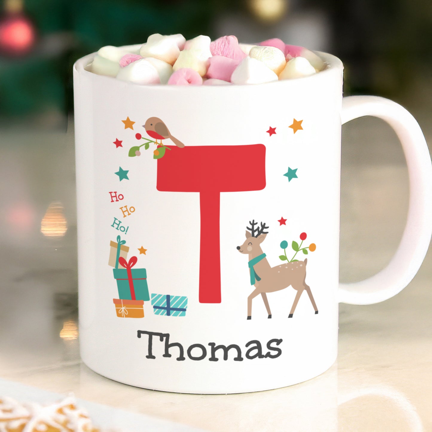 White mug with Christmas-themed design, including a reindeer, presents, and the name 'Thomas', filled with hot chocolate and marshmallows.