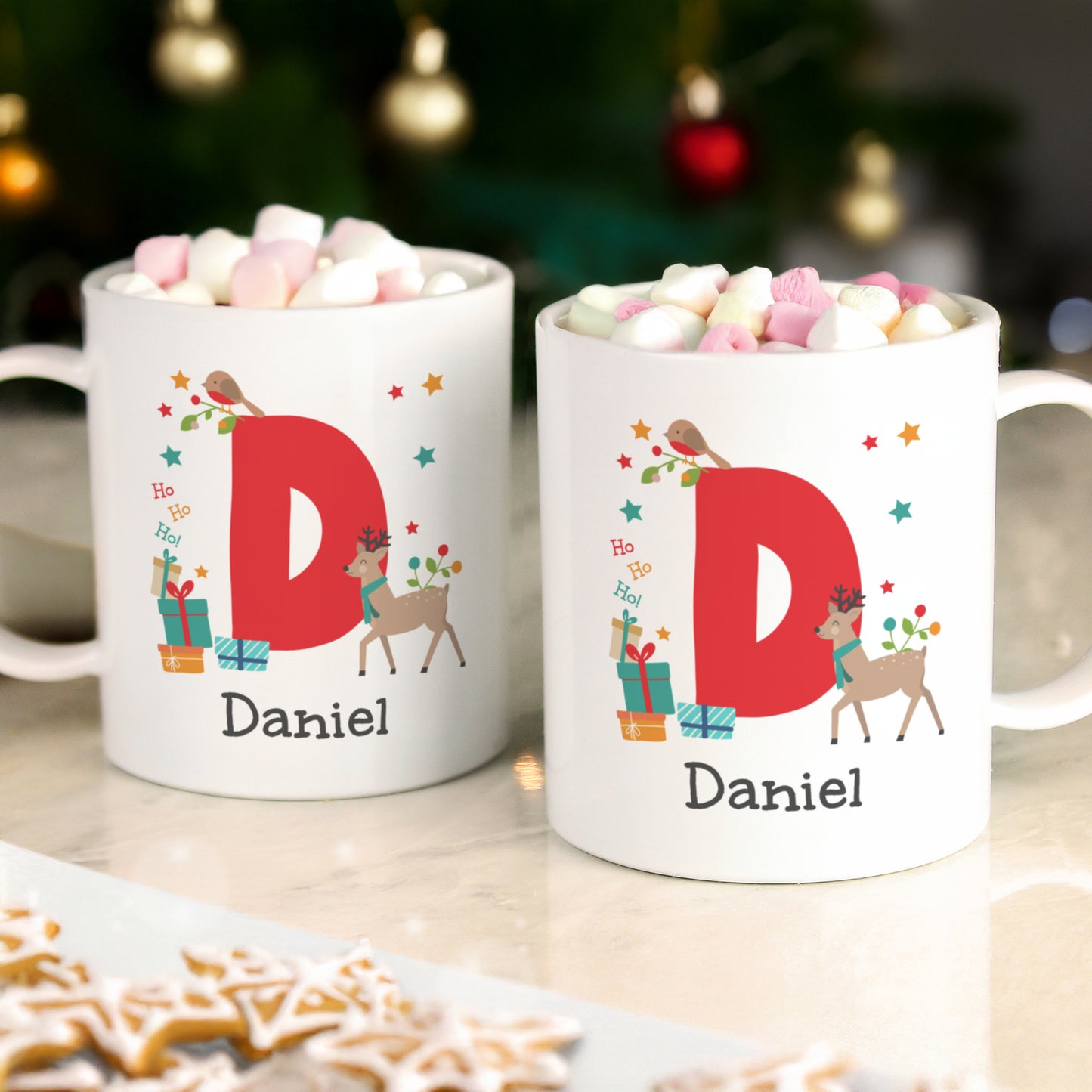 Two mugs with 'D' and 'Daniel' on a table with hot chocolate and cookies, Christmas tree in the background.