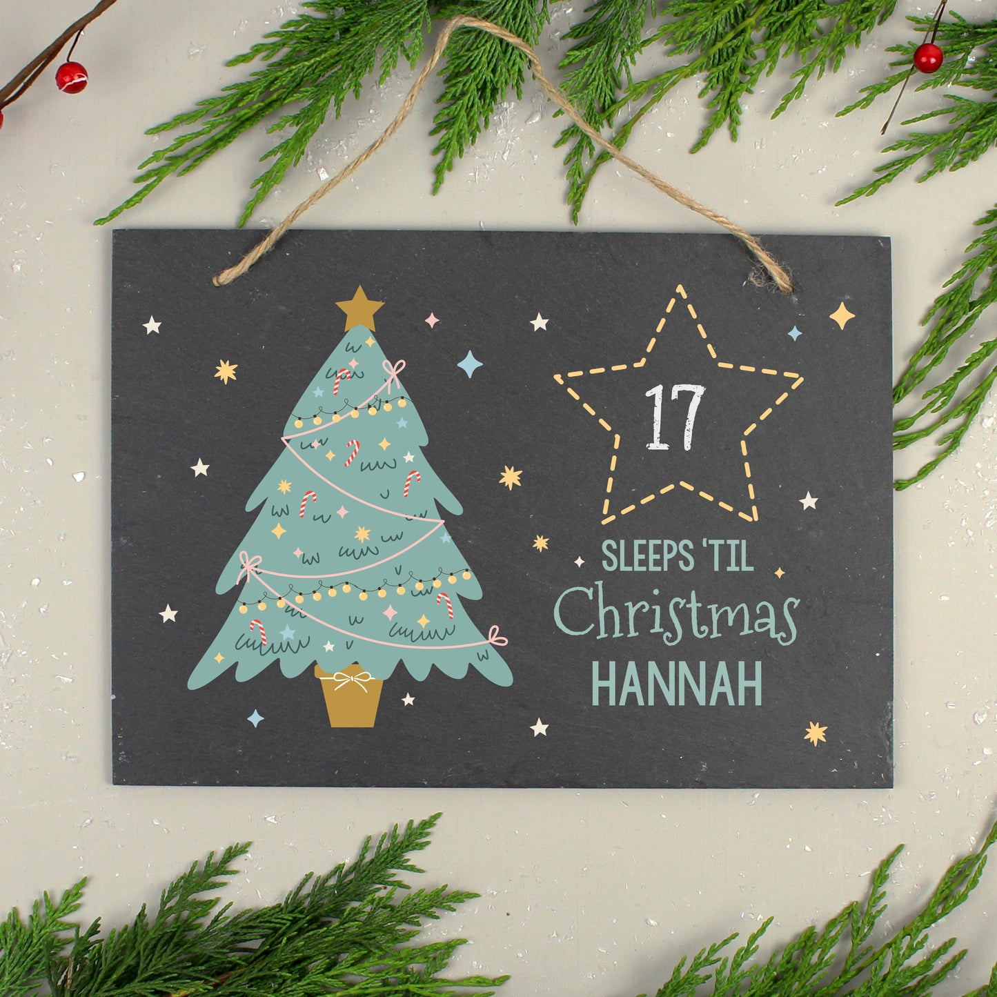 Personalised Christmas-themed slate with tree illustration and '17 sleeps til Christmas Hannah' text, surrounded by greenery.