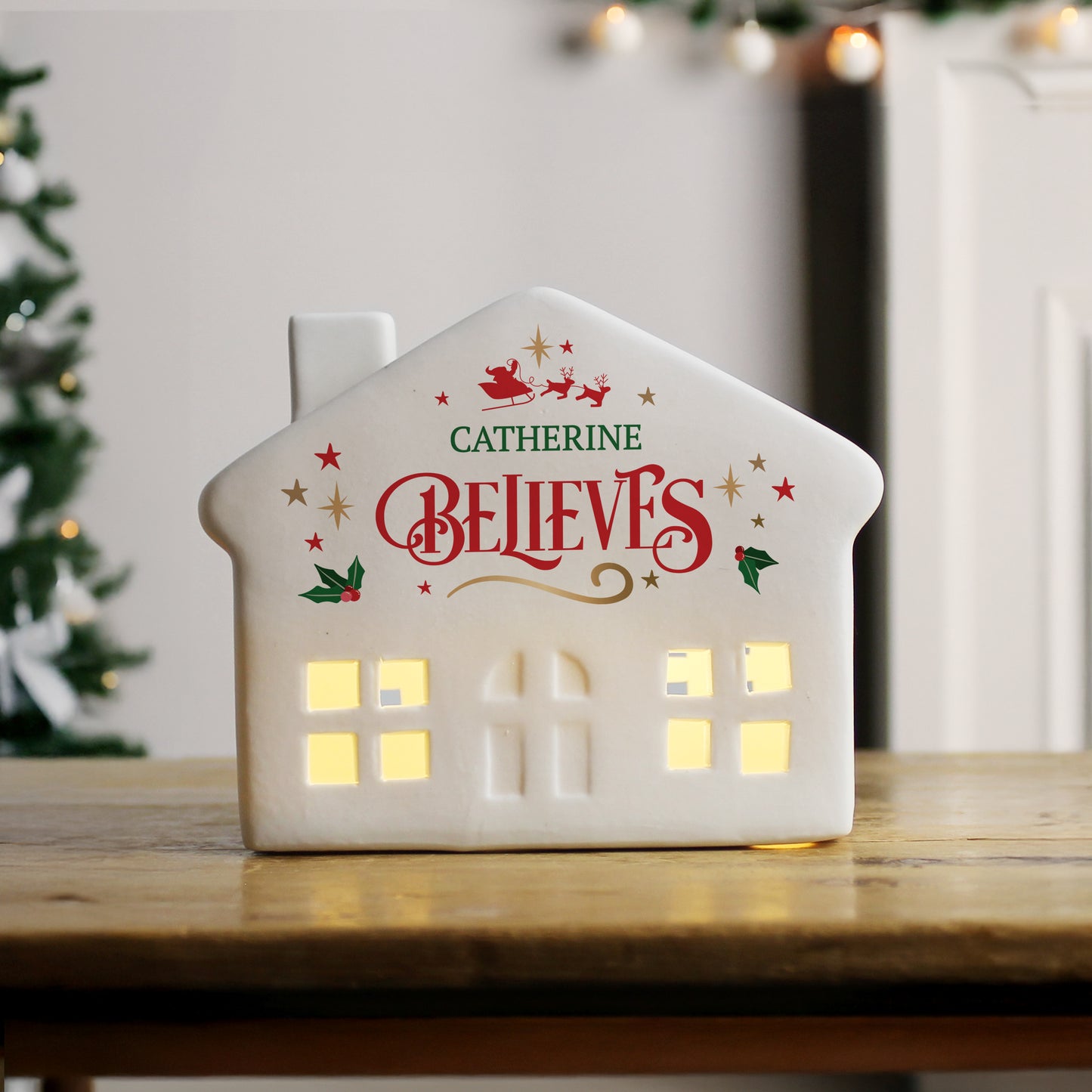 Decorative house-shaped light with 'Catherine Believes' text on a wooden surface with a Christmas tree in the background.