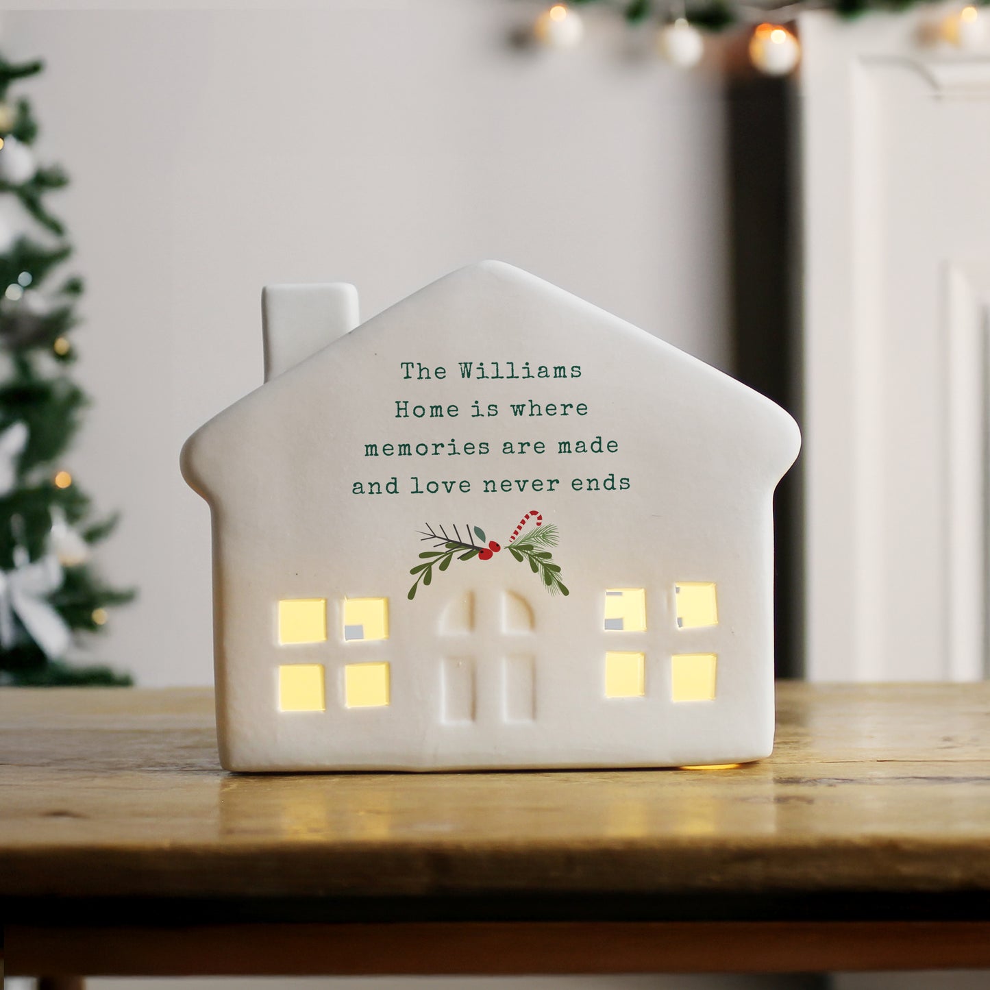 House-shaped decorative light with personalized message on a wooden surface with a blurred festive background.