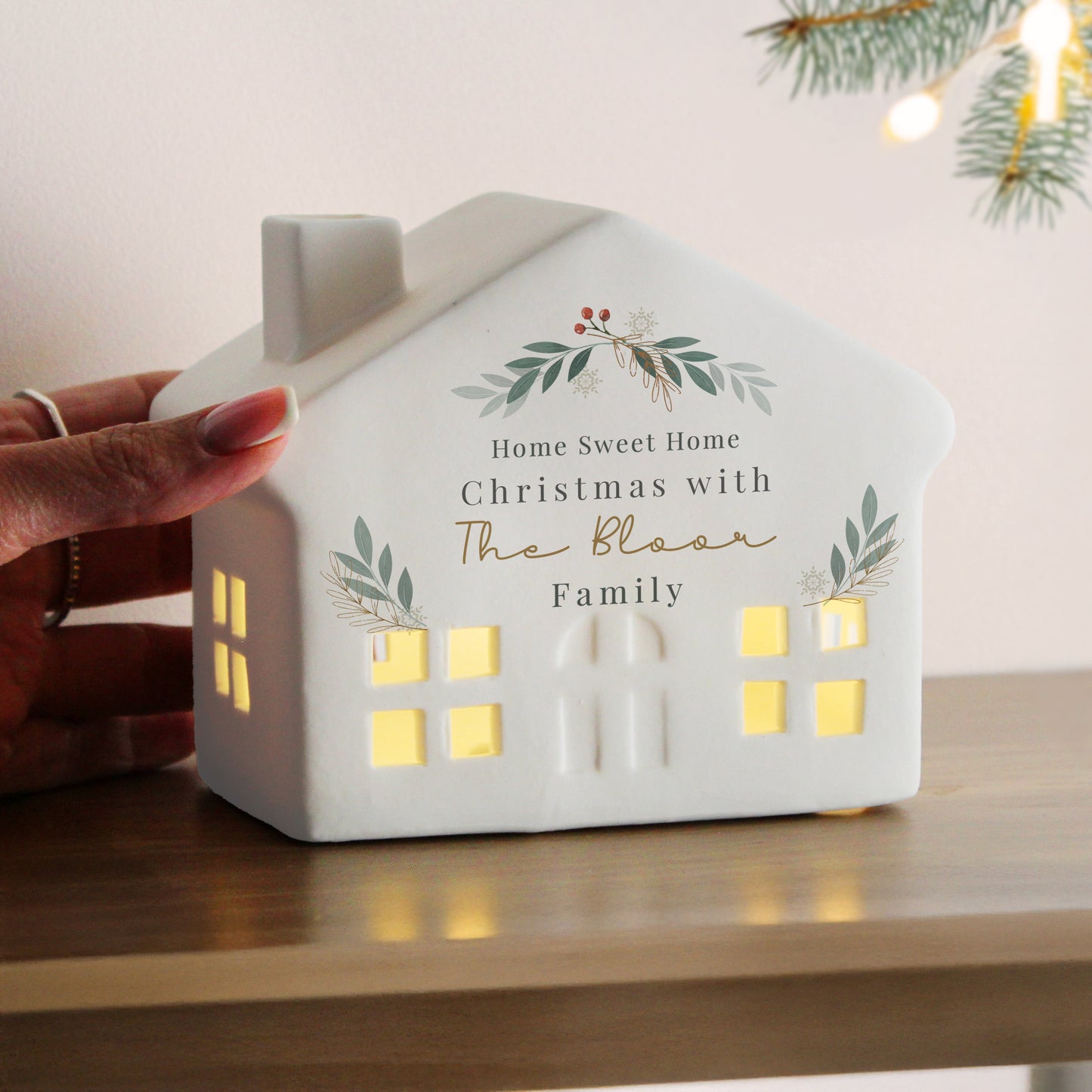 White house-shaped lamp with decorative text and greenery, held by a hand on a wooden surface.