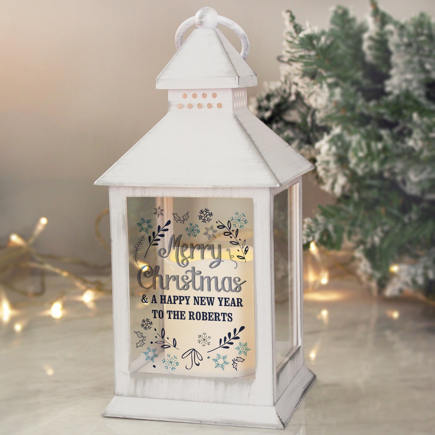White lantern with 'Merry Christmas & A Happy New Year' text in a decorative frame, with blurred Christmas tree and lights in the background.
