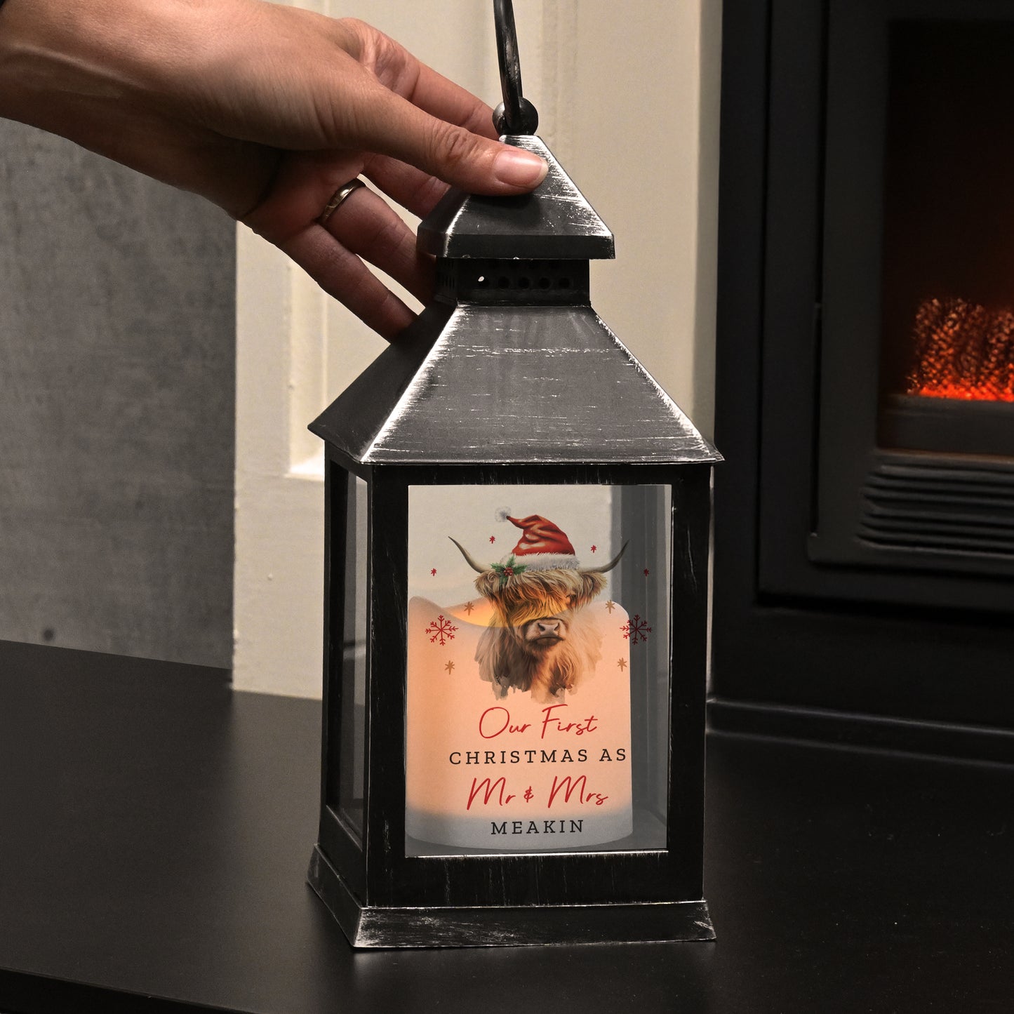 Black lantern with a festive card inside, held by a hand in front of a fireplace.
